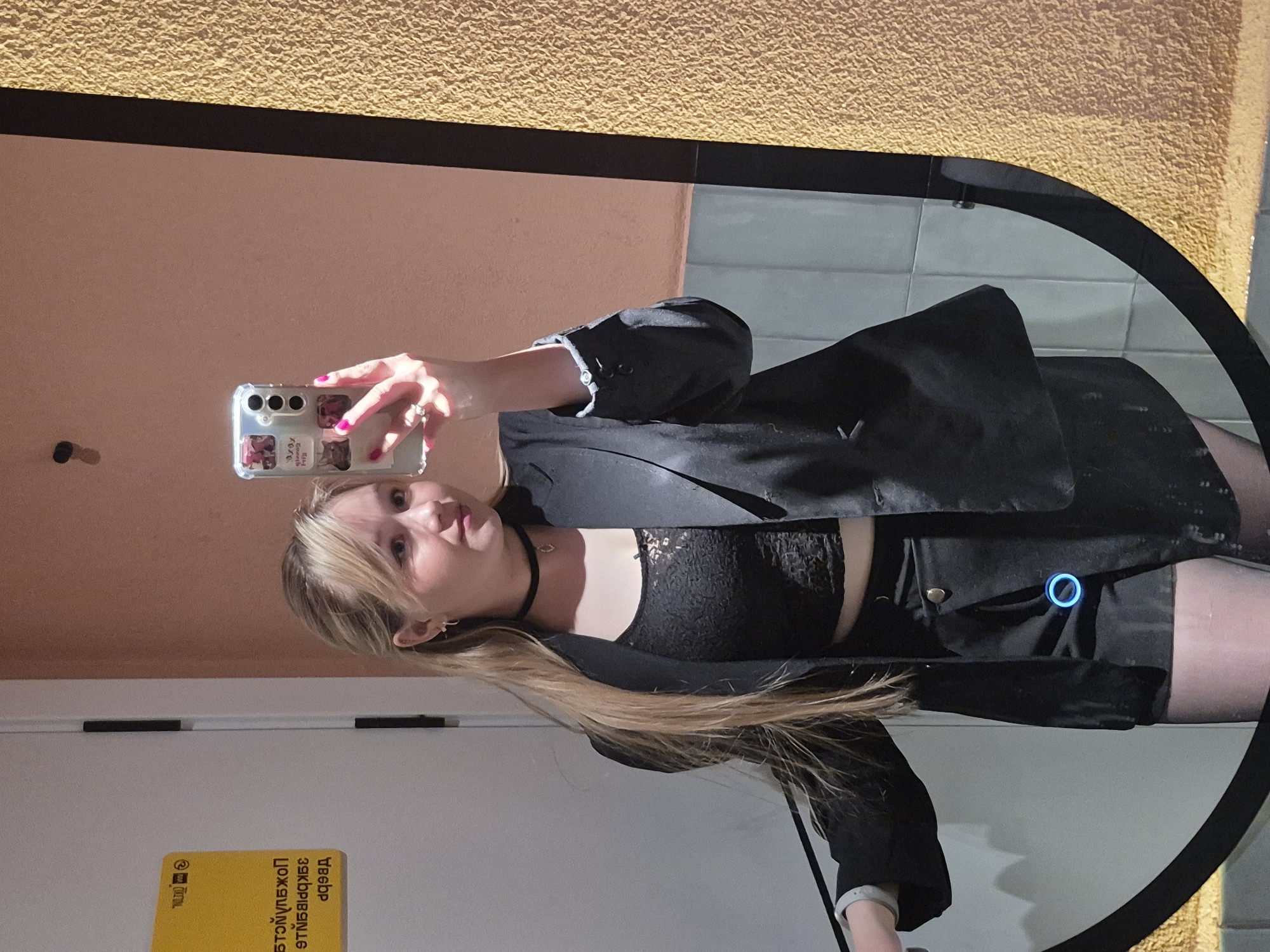 A young woman with blonde hair is taking a selfie in a mirror. She's wearing a black blazer over a black lace top and a black skirt or shorts. She has a black choker around her neck. She is holding a smartphone with a clear case decorated with stickers.
The mirror appears to be mounted on a wall in a public space. The walls are painted in different colors, including peach and yellow. The mirror's frame is black and somewhat circular. A door and a yellow sign with text in Russian are visible in the background. The location is Chaikovsky, Russia.