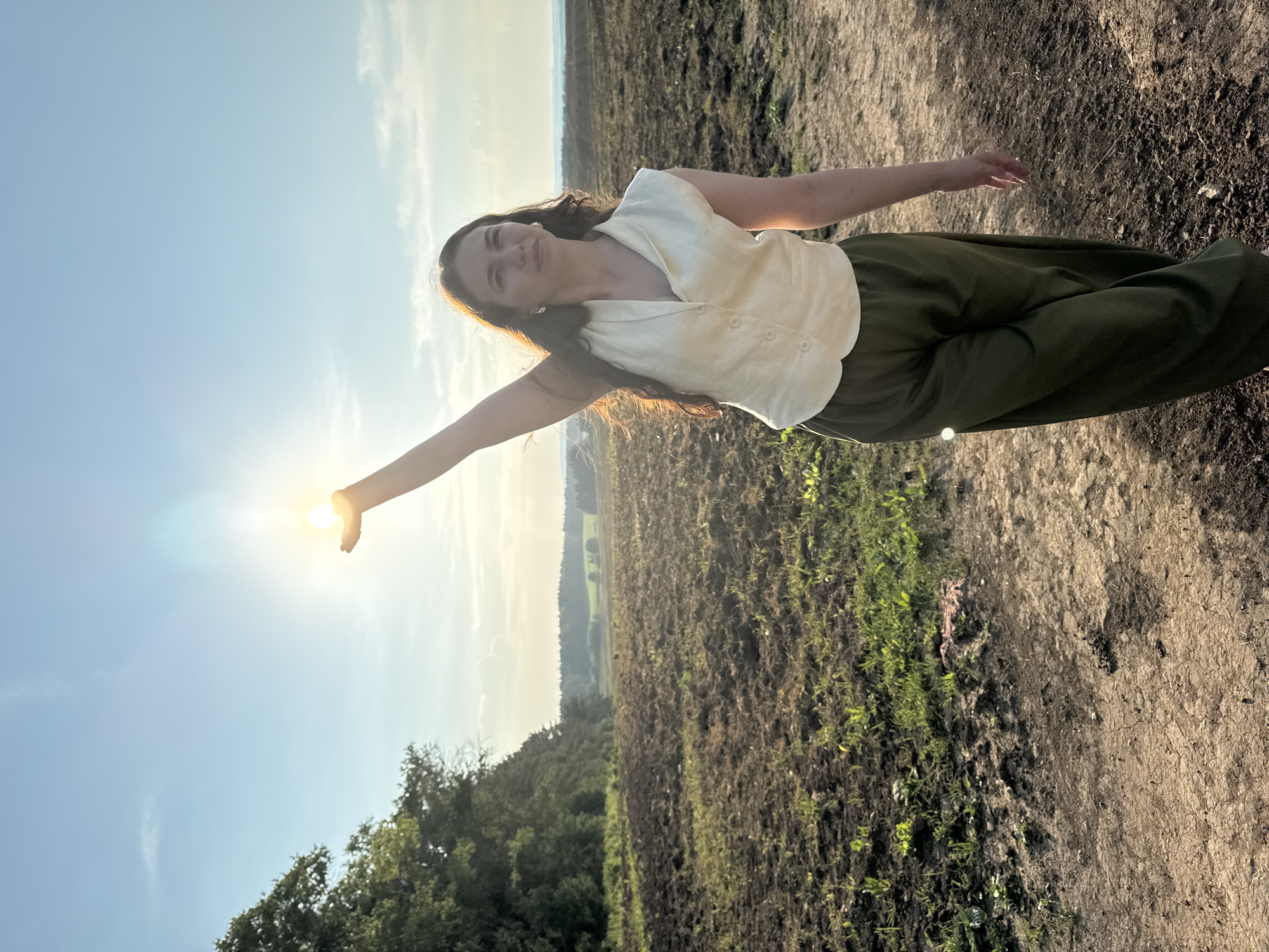 A young woman stands in a plowed field under a bright sky. She is wearing a cream-colored vest over a light-colored top and olive green pants. Her right arm is extended upwards, holding the sun in her cupped hand, creating a lens flare. The field is brown and dusty with some patches of green grass. In the background, there is a line of trees and a distant landscape under a hazy sky. The image appears to be taken at sunset. The location is identified as Lukintsy, Russia.