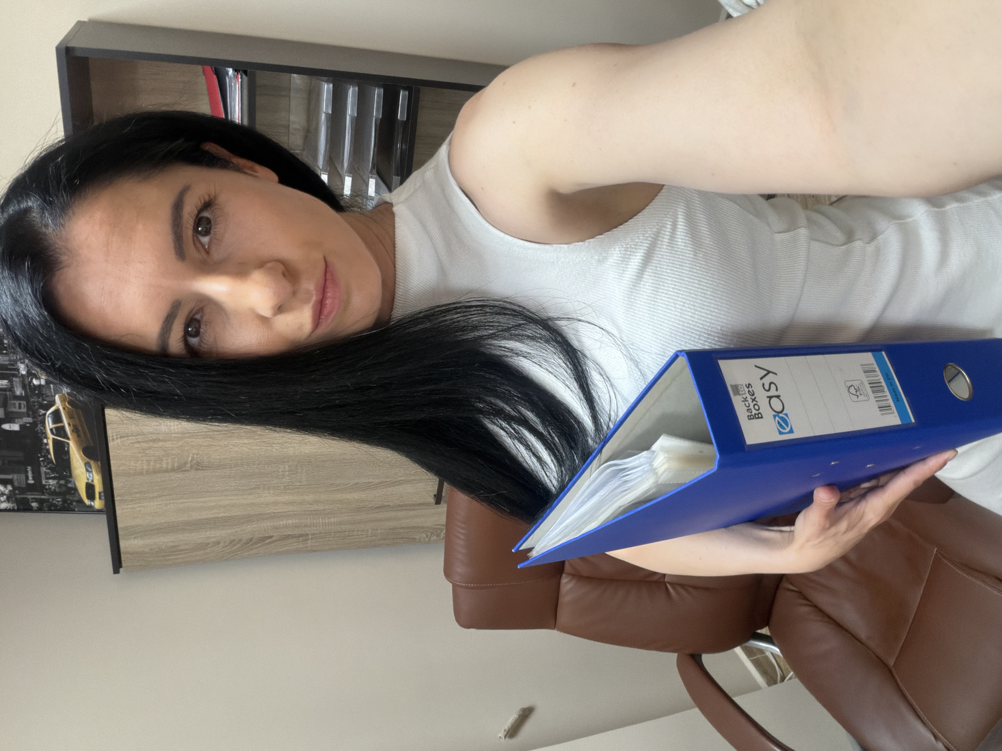 A woman with long, dark hair is holding a blue binder with the label "easy" on it. She is wearing a white ribbed tank top. She is sitting in a brown leather chair in what appears to be an office. Behind her, there is a light wood desk and a shelf unit with various items. A picture with a yellow taxi is seen behind the desk. There is no indication that this image was taken in Pleven, Bulgaria.