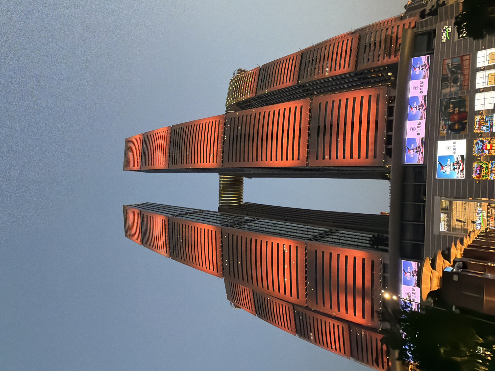 The image displays the Raffles City complex in Yuzhong District, Chongqing, China, captured during twilight or early evening. The central subjects are two massive, modern skyscrapers, which appear to lean inward as they ascend, forming a V-shape. A prominent, enclosed skybridge, also illuminated, connects these two towers at a high elevation.
Both skyscrapers are extensively lit with a vibrant orange-red light, which emanates from within or behind their structured facades. These facades feature numerous horizontal, slat-like or louvered panels that run the length of the buildings. Small, bright, scattered specks of light are visible across the red-illuminated surfaces of the towers and the skybridge, suggesting individual lighting elements or reflective accents.
At the base of the towers, a multi-story commercial structure is integrated. This podium level features several large digital display screens. Multiple screens prominently display an advertisement featuring the English word "Ultra" and Chinese characters that read "泰兰斯" (Tàilánsī). Another section of the building bears a white sign with the text "Raffles City" and additional Chinese characters. Below these signs, shopfronts and windows are visible, some displaying internal lighting or further promotional material. The sky overhead is a clear, muted blue, transitioning to a slightly darker hue towards the top of the frame, characteristic of dusk. No distinct human figures are individually discernible in the scene.