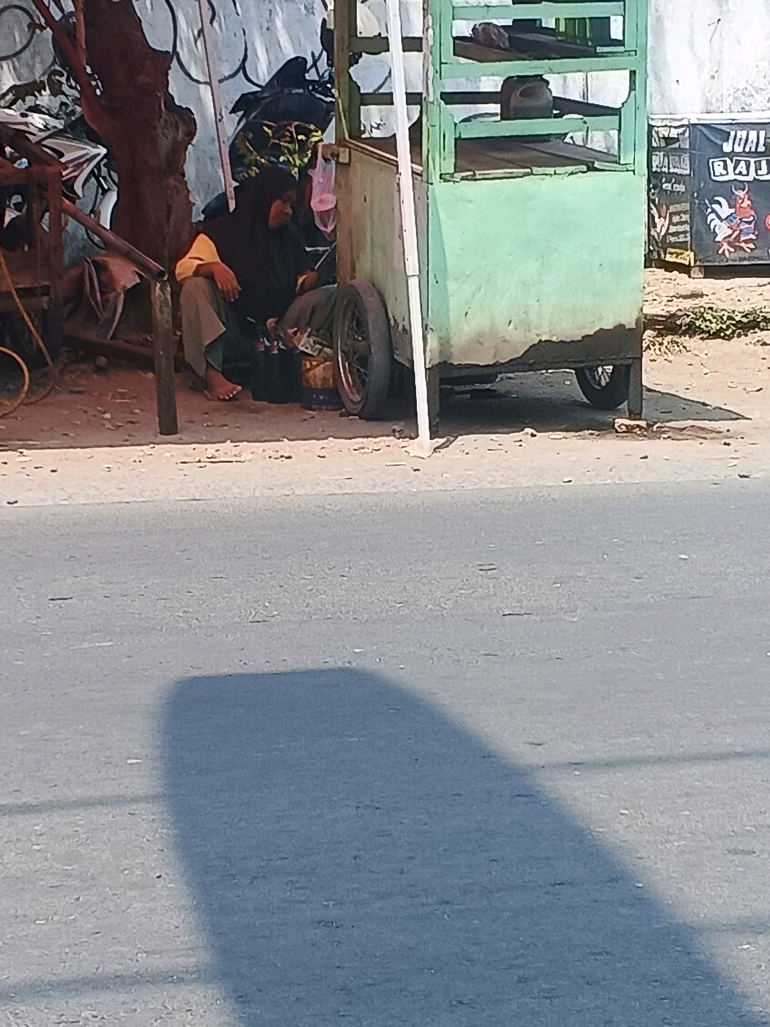 A person wearing a black hijab and grey pants is sitting on the ground next to a food cart in Taman, Indonesia. The food cart is green and has multiple shelves. A bicycle is partially visible behind the person. The person appears to be resting or waiting, with one foot bare. The ground is a mixture of dirt and pavement, and a street is visible in the foreground with a shadow cast across it. A sign with text and an illustration is attached to a structure behind the food cart.
