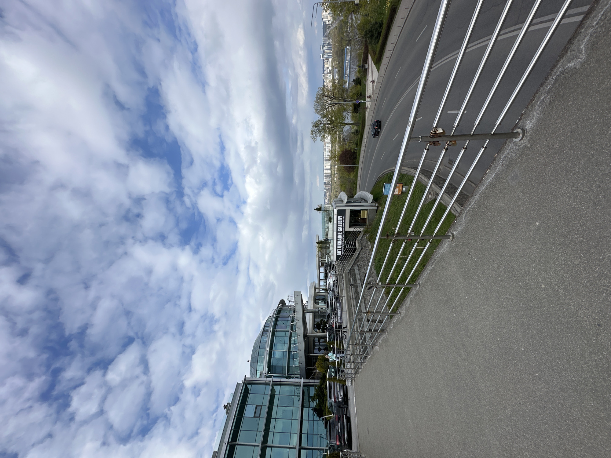 The image captures an elevated daytime view of an urban landscape in Kyiv, Ukraine, under a sky with scattered white and grey clouds and patches of blue.
In the foreground, a textured grey asphalt surface, likely a pedestrian path or bridge, is bordered by a modern metal railing. This railing consists of several parallel horizontal bars supported by vertical posts.
Beyond the railing, a multi-lane asphalt road curves through the scene, marked with white lines. A black car is visible on the road, driving away from the viewer. Adjacent to the road is a grassy area with trees, some of which are showing new green leaves, suggesting spring.
The midground features several modern buildings, predominantly constructed with glass facades. One prominent building has a distinctive curved, multi-story glass design. Below the main road, a smaller structure or part of a building clearly displays the text "ART UKRAINE GALLERY." Near this sign, a statue or sculpture is visible. A person wearing a light blue top is seen on a balcony or terrace of one of the glass buildings, appearing to wave or pose.
In the background, a wide body of water, likely the Dnieper River, stretches across the scene, with a bridge visible in the distance. Beyond the river, a dense cityscape with numerous buildings extends towards the horizon. The overall scene depicts a modern, active urban environment.