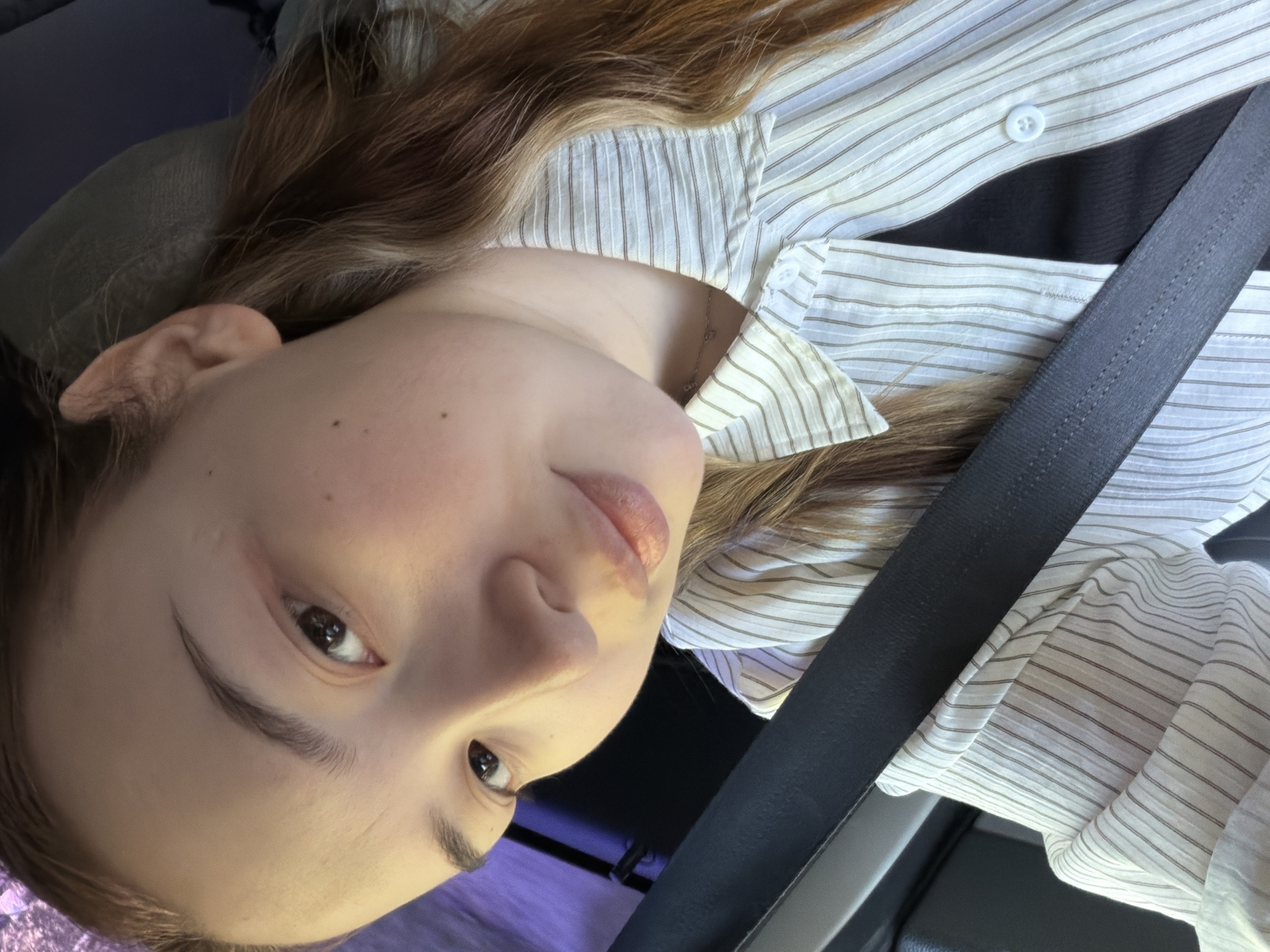 A close-up, tilted selfie shows a young woman with light brown hair. She is wearing a light-colored striped shirt and a black seatbelt. The setting appears to be the inside of a car. There is no specific information to indicate this photo was taken in Chaikovsky, Russia. No further details regarding the environment or location are discernible from the image.