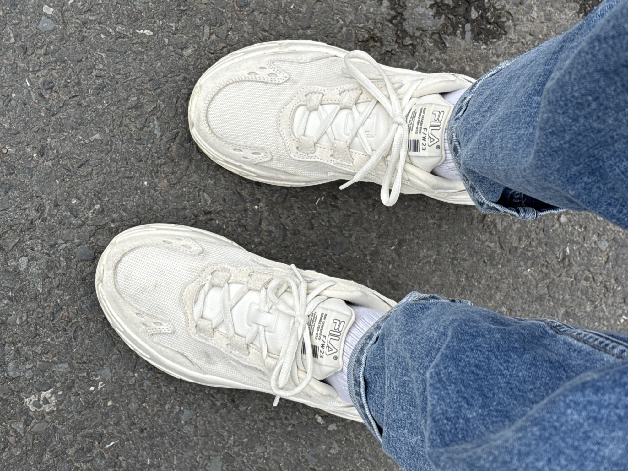 The image shows a view from above of a person's feet standing on a gray asphalt surface, likely a road or sidewalk. The person is wearing white FILA Ray Tracer Evo sneakers and blue jeans. Both shoes are tied. The tongues of the shoes are visible, as well as the labels, which contain "FILA" and "F/W 23 RAY TRACER EVD SRM02700-052". The person is wearing white socks. The location is not specified in the image.