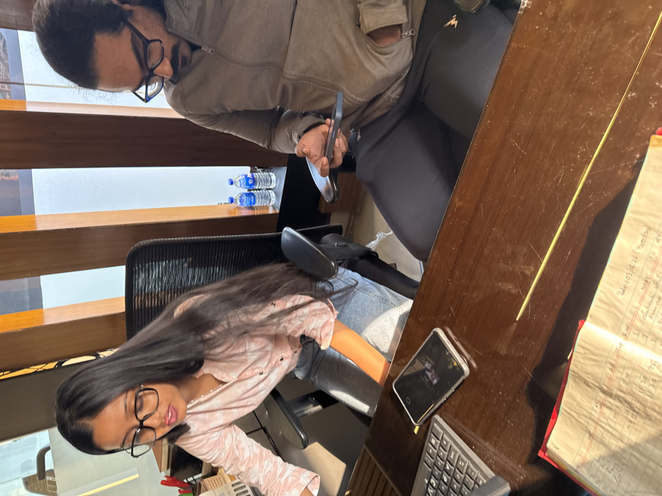 Two people are sitting at a desk. One person wears a brown jacket while looking at a phone. The second person is seated at the desk wearing a pink patterned shirt and glasses and working at a keyboard. A notebook and a cell phone rest on the desk. Water bottles are set on a shelf behind them. A wooden blind covers the background wall.