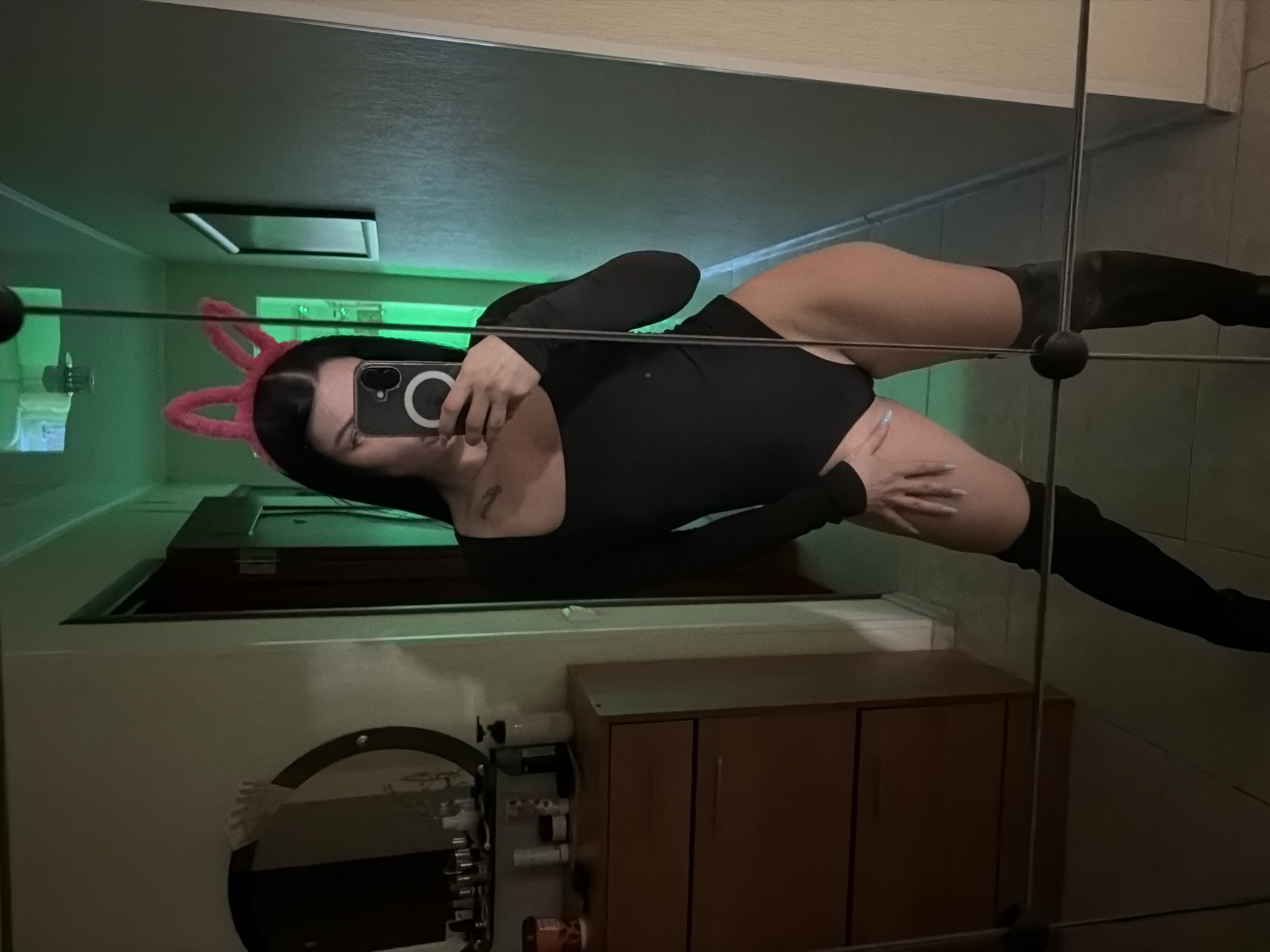 A woman is taking a mirror selfie in what appears to be a bedroom. She is wearing a black long-sleeved bodysuit, thigh-high black boots, and pink bunny ears. She is holding a phone in her right hand, which is angled to capture her reflection. To her left, there is a dresser with a round mirror above it, which reflects various items. The room is lit with green lighting. The flooring is tiled. There is a doorway visible behind the woman, leading to a hallway. The overall impression is of a personal space being used for self-portraits. The provided location of Ufa, Russia, is not visually verifiable from the image alone.