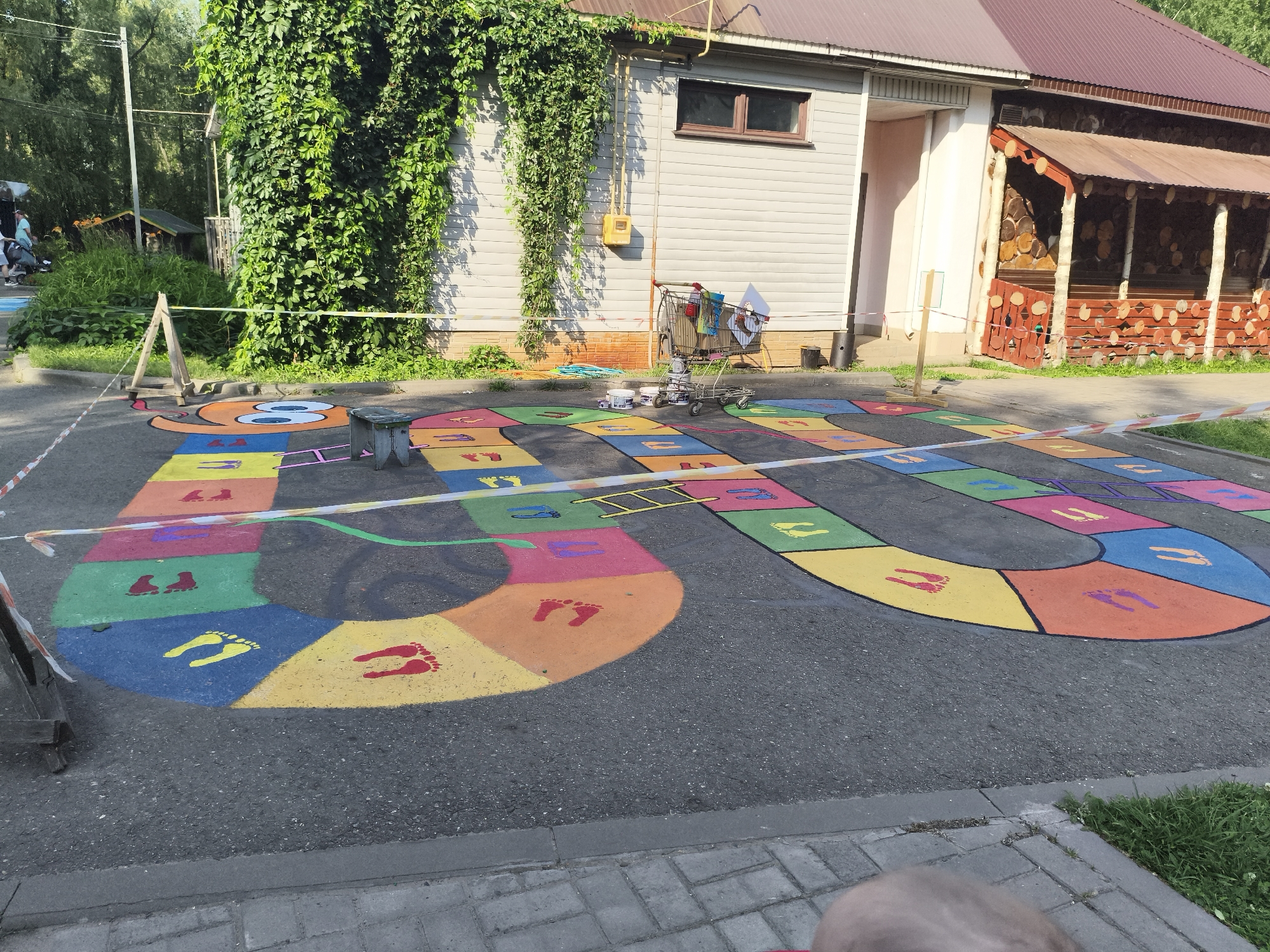 The image depicts an outdoor children's game painted on asphalt, resembling a hopscotch or board game. The game features a winding path of colorful squares, with footprints painted in various colors on some squares. A ladder is also visible on one of the squares, suggesting a "slide" or shortcut in the game. The game is cordoned off with caution tape. In the background, a building with light-colored siding and a dark roof is visible, partially covered in ivy. A shopping cart filled with items, possibly related to the painting of the game, stands near the building. The scene appears to be in a public outdoor area, possibly a park or playground, in Chaikovsky, Russia.