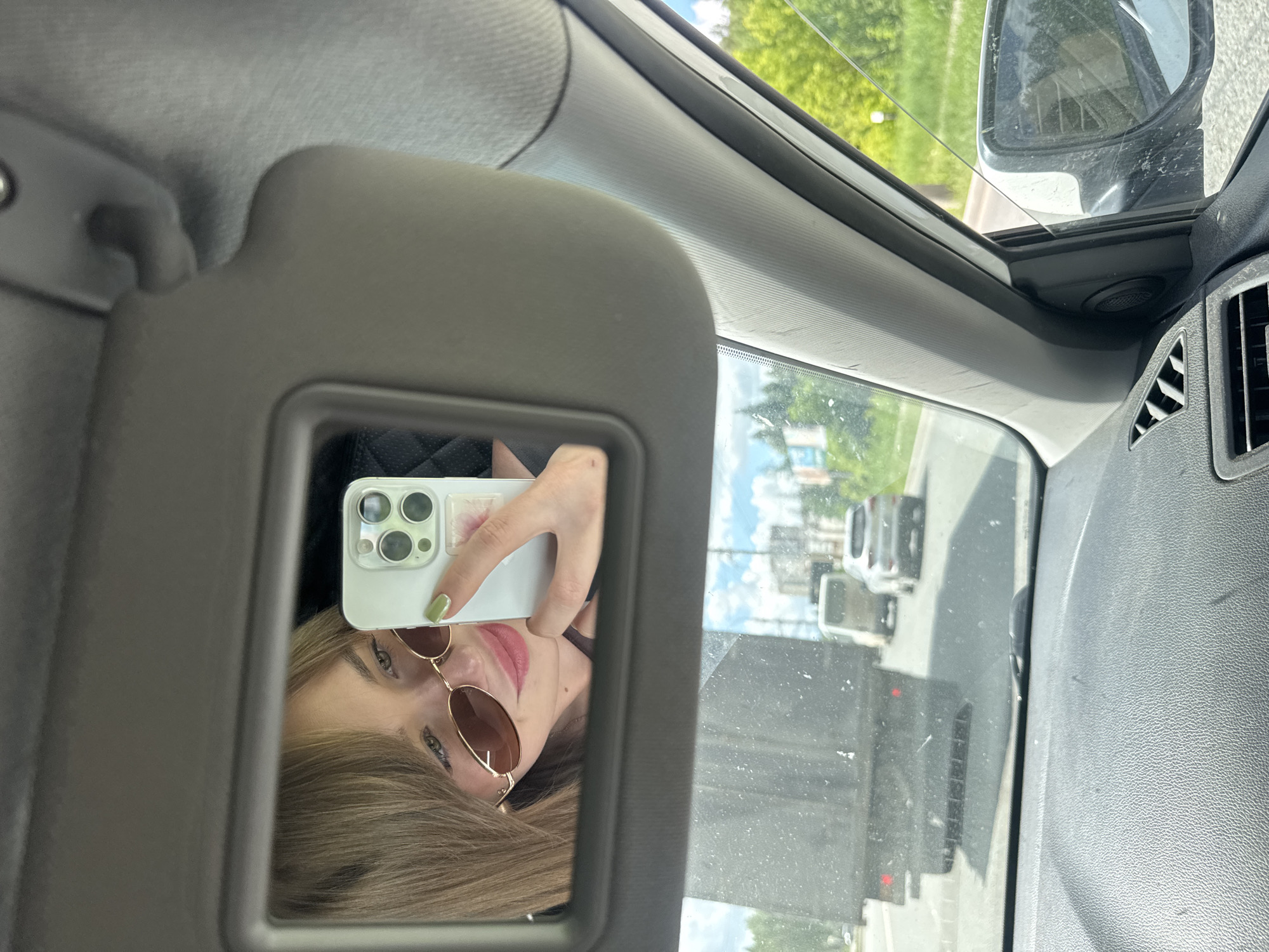 A woman is taking a selfie in the mirror of a sun visor inside a vehicle, likely a car. The woman is wearing sunglasses and has light-colored hair. She is holding a white smartphone, possibly an iPhone, with multiple lenses. The view outside the car window shows a road with other vehicles, including a truck and cars. The location might be Izhevsk, Russia, but there's nothing to confirm that detail.