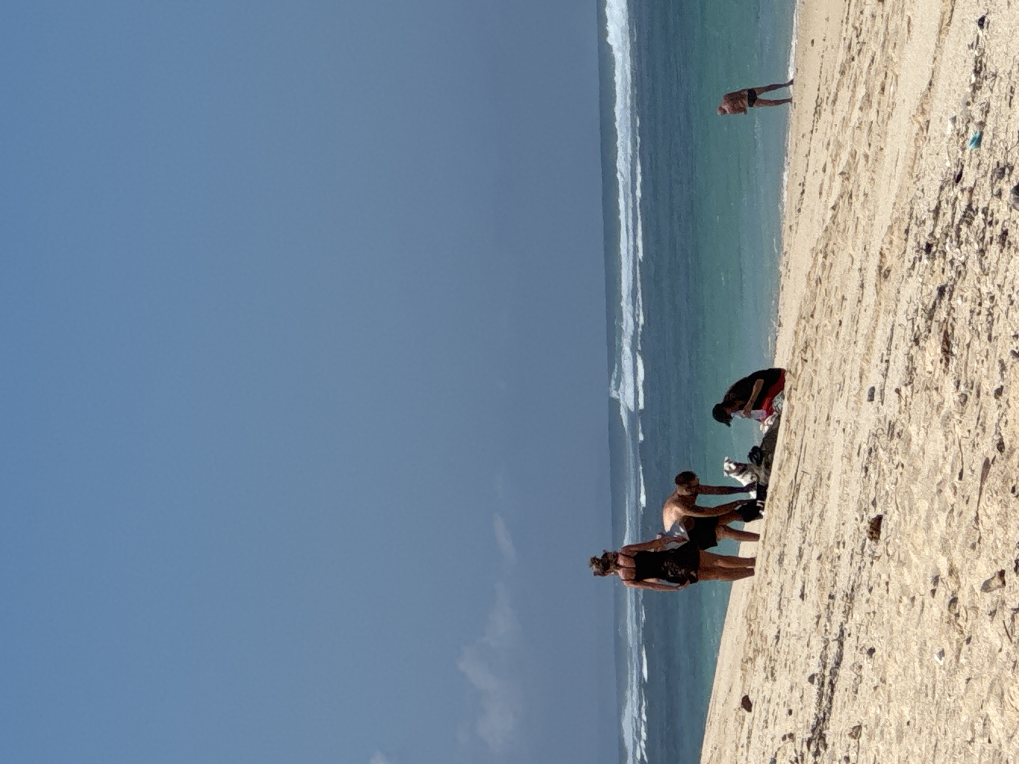 A group of people are on a sandy beach with the ocean in the background. The sky is a clear blue with some clouds towards the horizon. The ocean has waves breaking on the shore. In the foreground, there are a few scattered rocks and debris on the sand. A woman in a black dress and a man in swim trunks are crouched down, appearing to be interacting with a dog. Another person is seated on the sand further down the beach. On the right side of the image, a man in dark swim trunks is standing near the water's edge. The scene suggests a casual day at the beach in Kuta Selatan, Indonesia.
