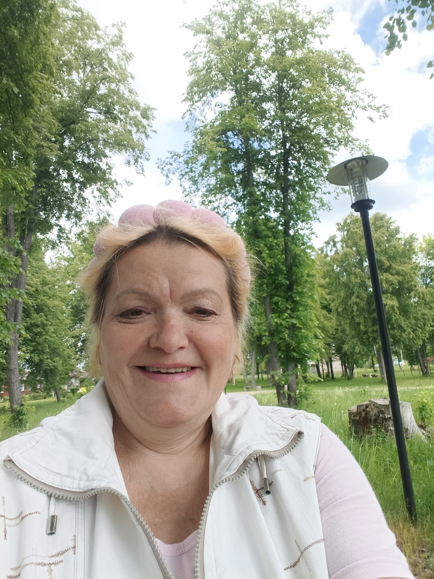 A cheerful, middle-aged woman with blonde hair, styled with pink rollers, smiles warmly directly at the camera in a vibrant outdoor setting. She is wearing a light-colored, possibly off-white, zipped sleeveless vest over a pale pink long-sleeved shirt. Her expression conveys happiness and contentment.
The background is dominated by lush green deciduous trees with bright foliage, suggesting a park or a spacious green area. Patches of blue sky with white clouds are visible through the tree canopy, indicating a pleasant, likely sunny or partly cloudy daytime. On the right side of the image, a tall black lamp post with a white fixture stands vertically. Further back, green grass stretches across the ground, and faint hints of other park elements like tree stumps can be seen. The woman appears to be taking a selfie or being photographed in this serene environment. The scene is located in Красный Маяк, Russia. There is no visible text in the image.