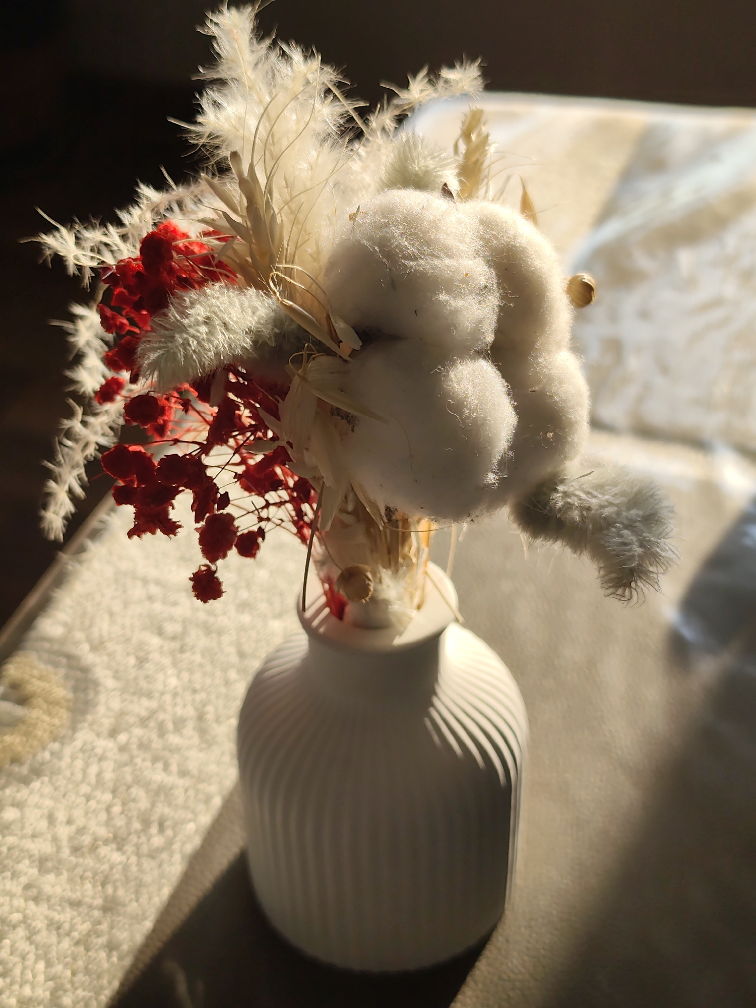 A still life photograph shows a white vase filled with dried flowers. The vase is white and round with vertical ridges. The flowers are a mix of white, beige, and red elements, including cotton boll. The arrangement appears to be placed on a beige or light-colored surface, possibly a table or couch. The scene is illuminated by warm, natural light.
The location in the city of Нижний Пальник, Russia cannot be verified as part of the current image.