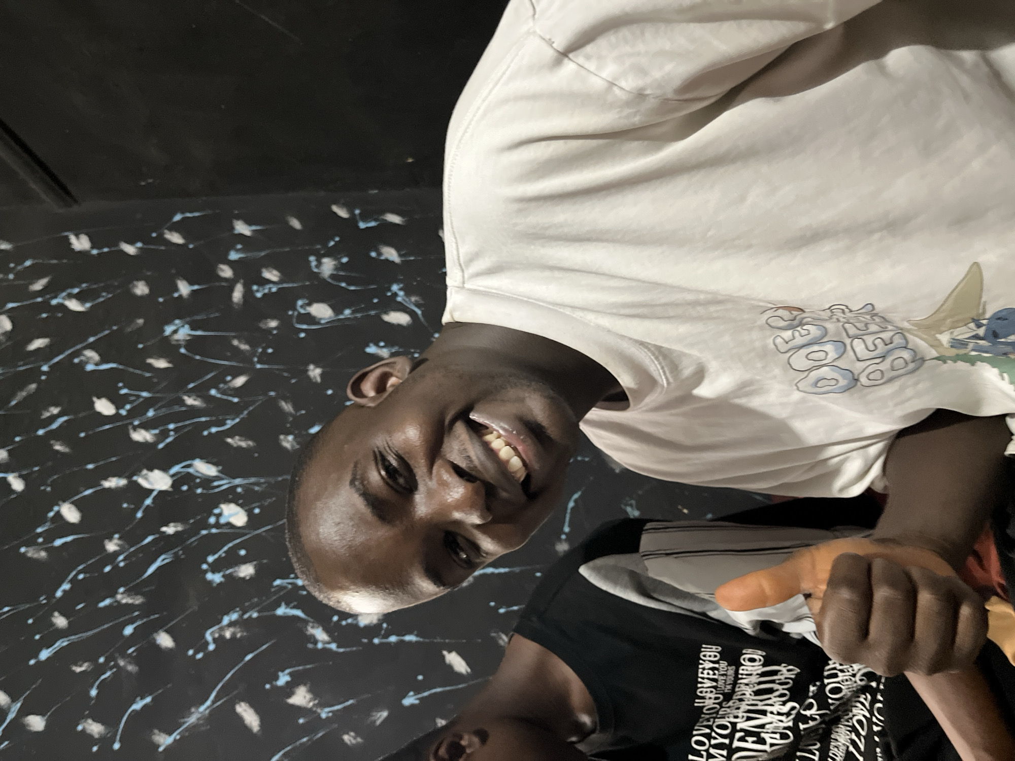 The image shows two young Black men in what appears to be a dimly lit indoor setting with a black wall decorated with blue and white paint splatters. The man in the foreground, facing the camera, is smiling broadly and giving a thumbs-up. He is wearing a white t-shirt with a graphic design that is partially visible. To his left, another man is seated, wearing a black t-shirt with white lettering. The overall impression is of a casual, friendly gathering. Based on the context provided, this scene is located in Jos, Nigeria.