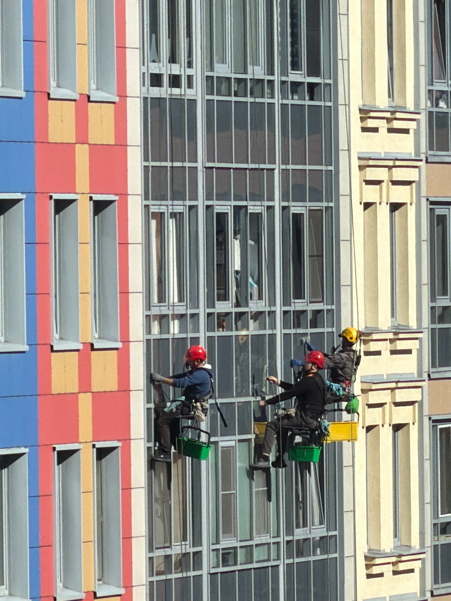 The image shows a colorful, modern building with many windows. The facade of the building is divided into vertical sections of blue, red, and yellow. There are three window cleaners suspended by ropes on the side of the building. Two of the cleaners are wearing red helmets, and one is wearing a yellow helmet. They are all wearing harnesses and carrying equipment. The cleaner on the left appears to be actively cleaning a window. The other two cleaners are seated in suspended platforms. The building is made of glass and metal, with cream-colored accents. The scene is set in a city, likely Saint Petersburg, Russia, given the architectural style.