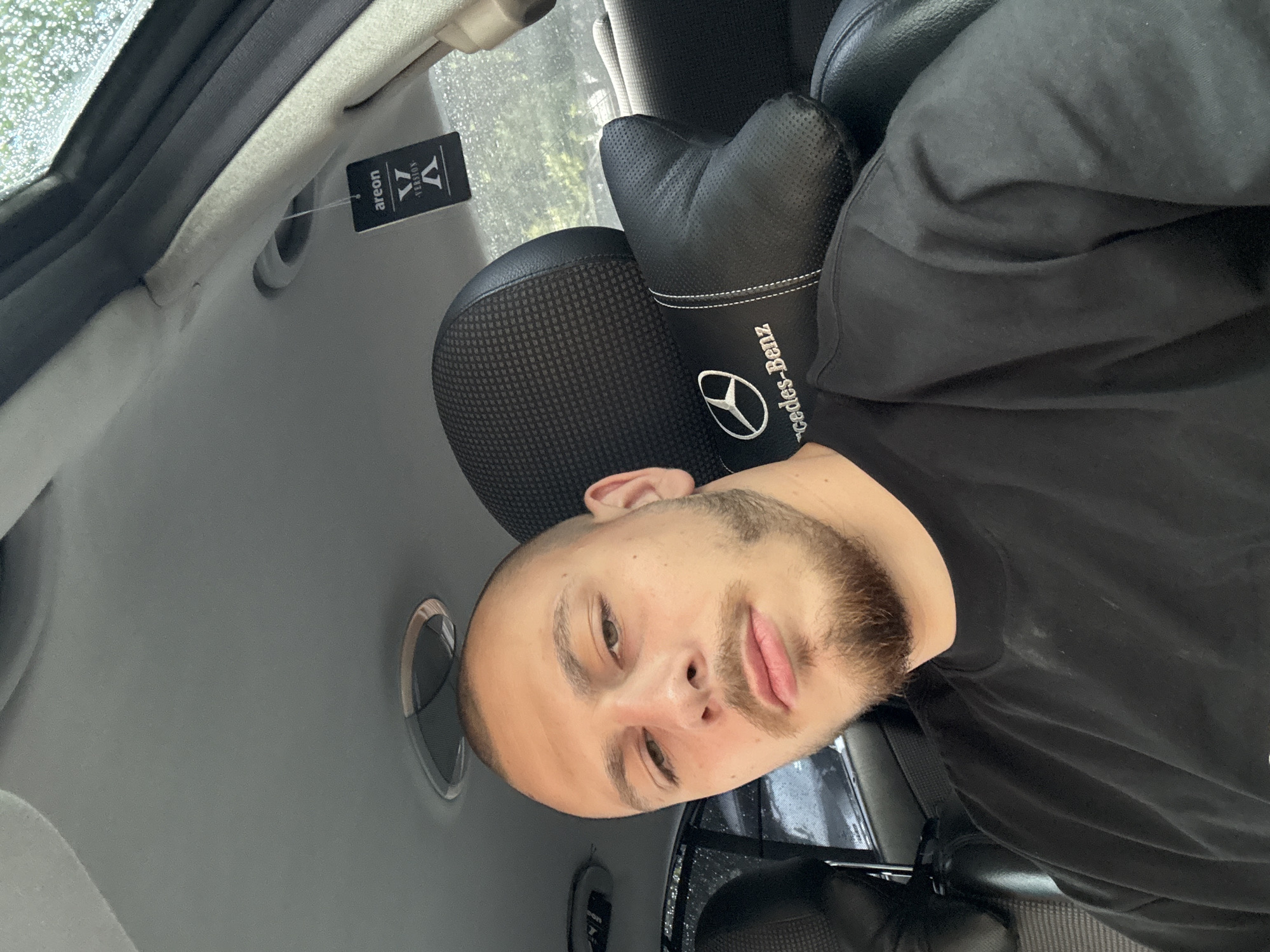 The image depicts a young male seated in the front passenger seat of a vehicle, taking a selfie. He has a shaved head and a short, light brown beard and mustache. He is wearing a plain black crew-neck shirt. The car's interior features a black seat with a mesh-patterned headrest and a black, perforated leather-like neck pillow with white stitching, embroidered with a Mercedes-Benz logo and text. A black rectangular "areon VERSION X" air freshener hangs from an overhead grab handle. The car's headliner is grey. Through the window, raindrops are visible, suggesting rain, with blurred green foliage in the background. The scene is located in the city of Stara Zagora, Bulgaria.