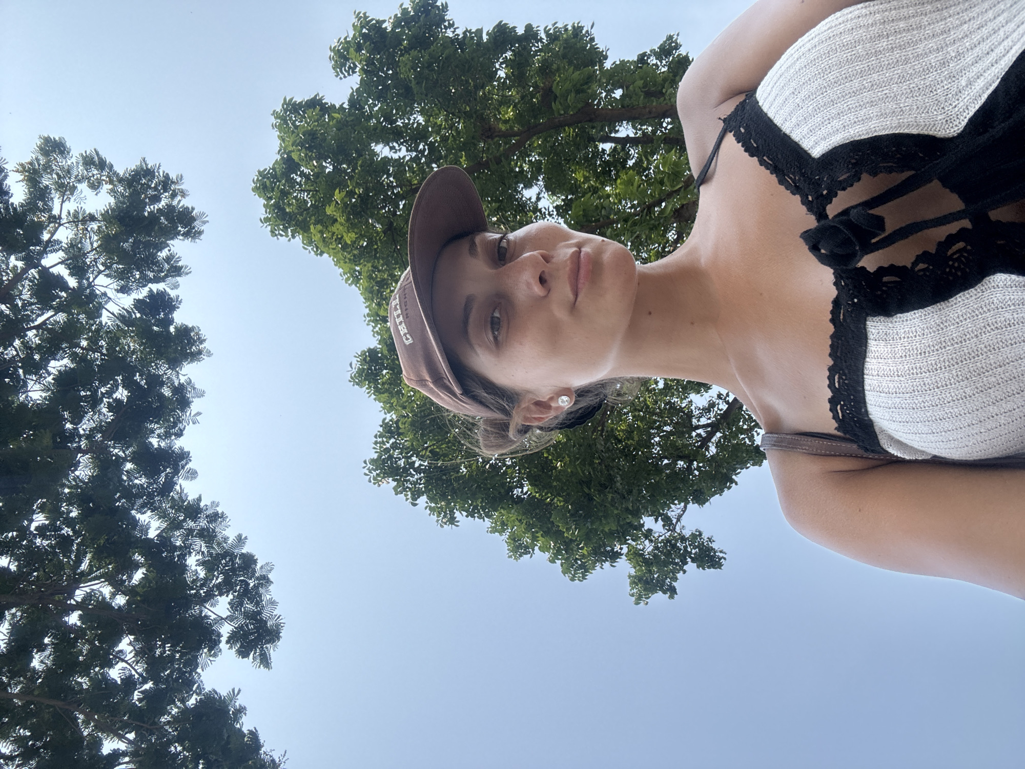 A woman is depicted in a selfie-style photograph taken from a low angle, with the camera pointing upwards towards her. She wears a brown baseball cap, a white knitted top with black lace detailing, and a single stud earring is visible. Her face is well-lit, and she appears to be looking directly at the camera. Behind her, two trees with lush green foliage are visible against a clear blue sky. It is not possible to determine if this is Đà Nẵng, Vietnam.