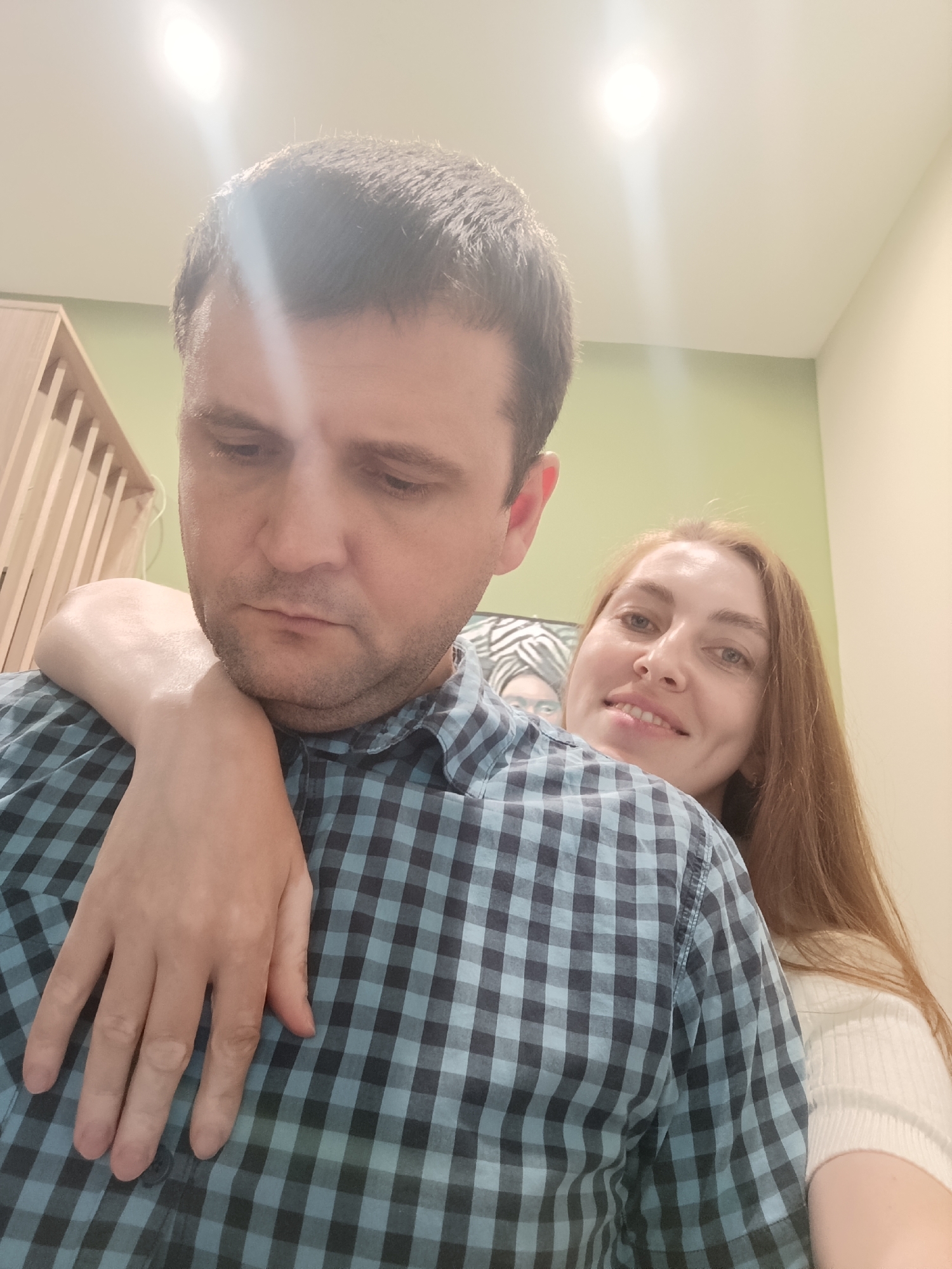 This is an indoor portrait captured in Moscow, Russia, featuring two individuals: a male and a female. The male is positioned in the foreground, his head and upper torso occupying the left and central parts of the frame. He has short, dark hair, light stubble, and his gaze is directed downwards. He is wearing a blue and dark blue plaid button-up shirt. The female is situated behind and to the right of the male, with her head visible over his left shoulder. She has long, reddish-brown hair, is smiling, and is looking directly at the camera. Her right arm is draped over the male's left shoulder, and her hand rests on his upper chest area. She is wearing a white ribbed long-sleeved top.
The background consists of light-colored walls; a light olive green wall is visible to the left and center, while a light beige wall is on the right. A partially visible wooden slatted structure is in the far left background. Behind the female, a portion of a framed image or artwork depicting a person with a patterned head covering is visible. The ceiling is white, featuring a bright, overhead light source positioned in the upper center-left, creating a significant lens flare and overexposure in that area.