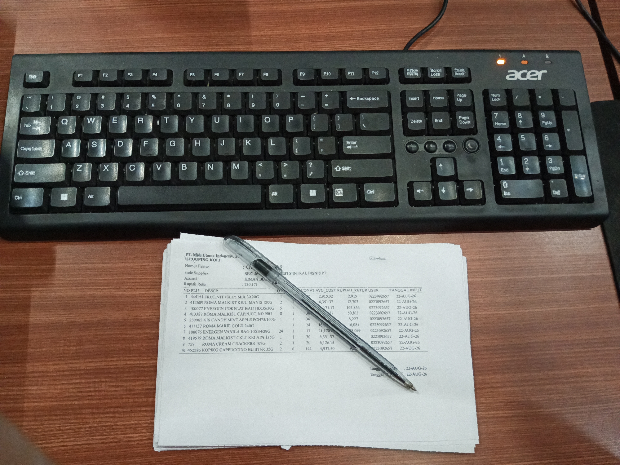 The image shows a black Acer keyboard and a stack of papers with text and a pen on a wooden desk. The papers appear to be a list of products with details like product number, description, quantity, price, and input date. The keyboard is a standard QWERTY layout with a numeric keypad on the right. The pen is a blue ballpoint pen. The wooden desk surface is visible. The scene is indoors, likely an office environment. There is no indication of the city of Makassar, Indonesia.
