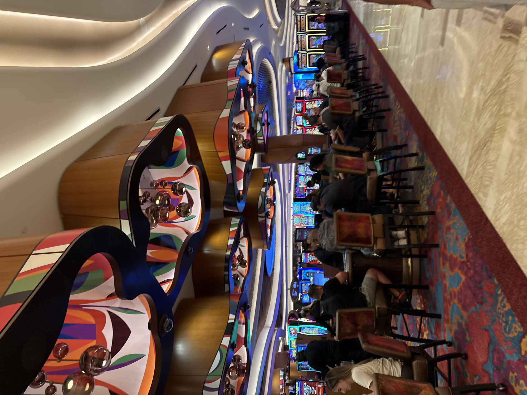 The image shows a scene inside the Seminole Hard Rock Hotel & Casino, located in the Hollywood Seminole Reservation, Florida, USA. Rows of slot machines line the side of the room, with numerous people seated and playing them. There are ornate decorative elements, including curved structures with colorful patterns and glass spheres, along the corridor. The floor is covered in a patterned carpet. The lighting is a combination of natural light and artificial illumination from the machines and overhead lights.