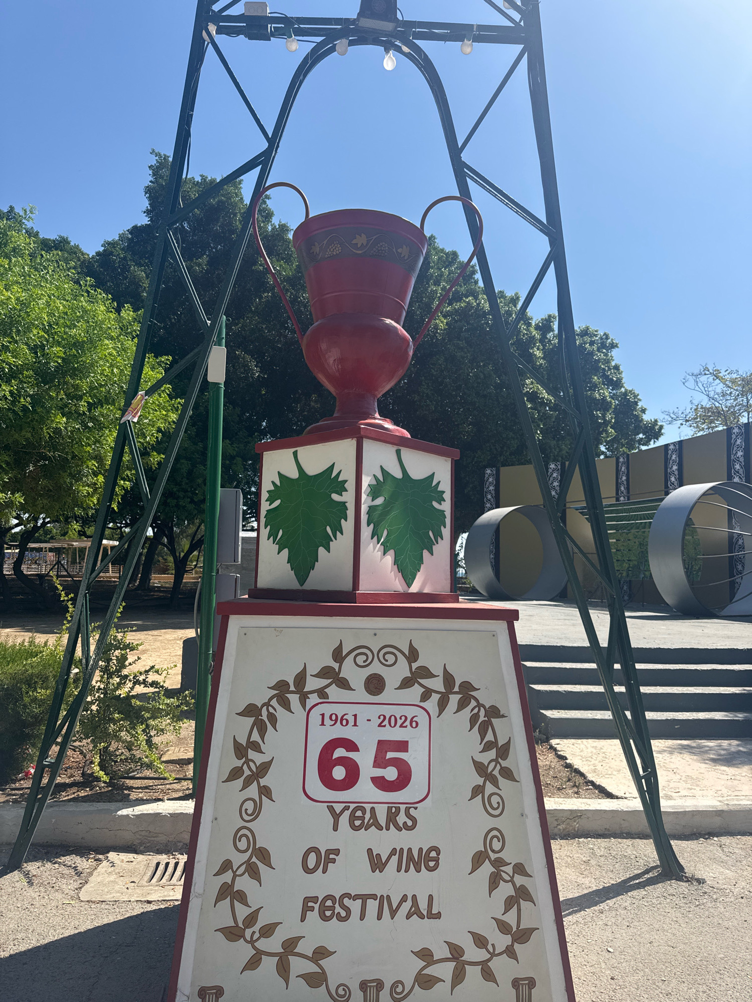 The image displays a large trophy-like centerpiece, painted red, atop a pedestal. The pedestal has white panels decorated with green grape leaves. Below this, a larger sign in a decorative frame reads "1961 - 2026 65 YEARS OF WINE FESTIVAL". The structure is set against a backdrop of trees and a clear blue sky. In the background, there are large, gray, cylindrical structures and a set of stairs. The scene appears to be outdoors, possibly in a public space or park. The trophy and sign suggest a celebration or event, specifically a wine festival. The presence of the trophy and decorative elements points towards a festive atmosphere. The overall impression is one of a celebratory display in an outdoor setting. The specific location in Limassol, Cyprus, is not explicitly indicated in the visual details of the image.