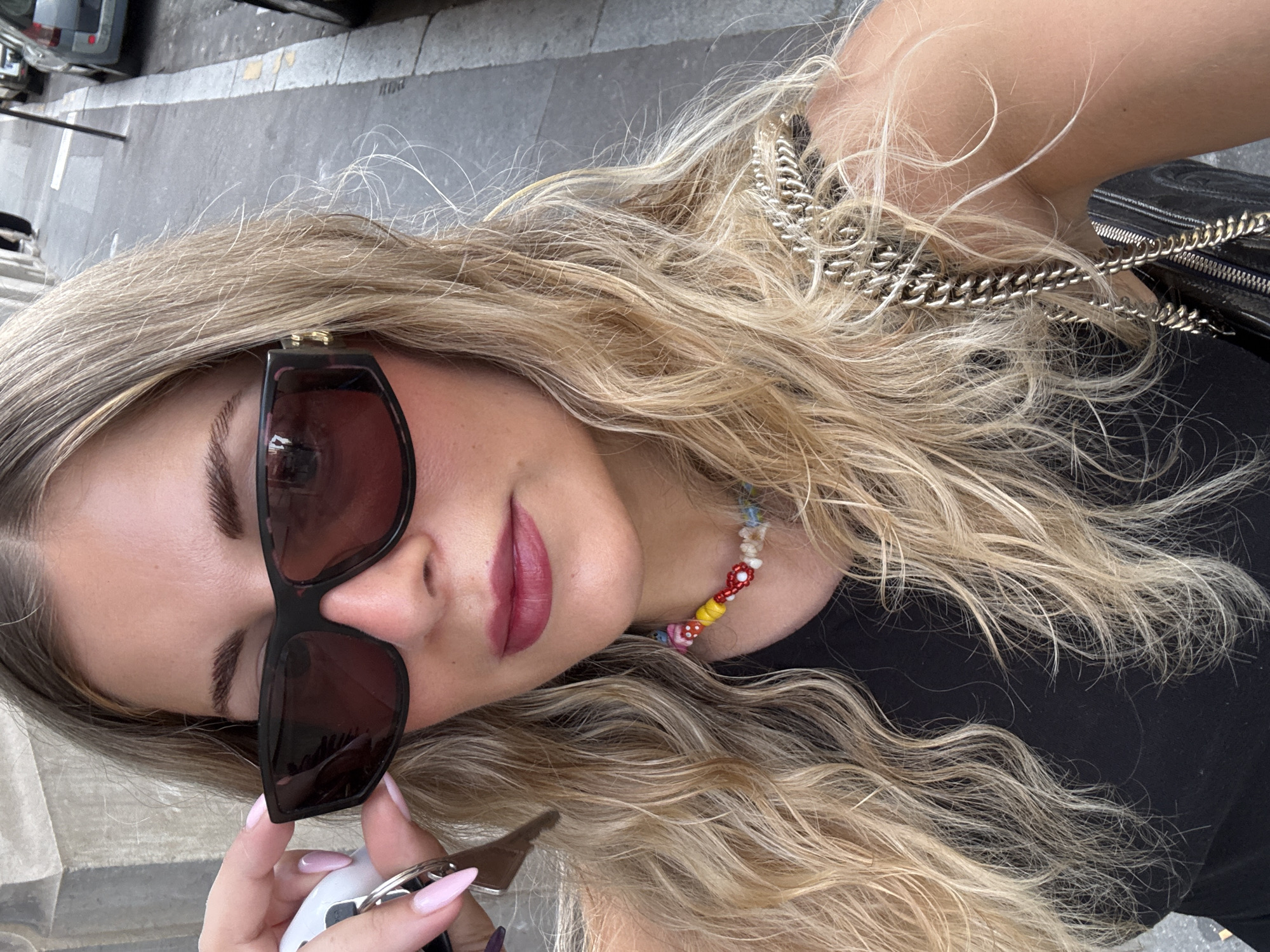 A tilted, close-up shot captures a person from a high angle, looking towards the camera. The subject has long, wavy, blonde hair that frames their face and extends around their shoulders. They wear large, dark-lensed sunglasses with prominent frames, obscuring their eyes. Their lips are adorned with a deep pink or plum-colored lipstick, and their eyebrows are well-defined.
Around their neck is a vibrant, colorful beaded necklace featuring various shapes, including floral and mushroom designs, alongside simple round beads. A silver chain strap, likely belonging to a handbag, is visible across their shoulder and intertwined with their hair. The person is wearing a black top. In their left hand, which has manicured light pink, pointed nails, they hold a set of keys, one of which appears to be a car key.
The background reveals an urban outdoor setting, with gray pavement or a sidewalk and a partially visible parked car in the upper left corner. The lighting suggests it is daytime. The image portrays a casual, stylish moment in what is identified as Paris, France. No visible text is present in the image.