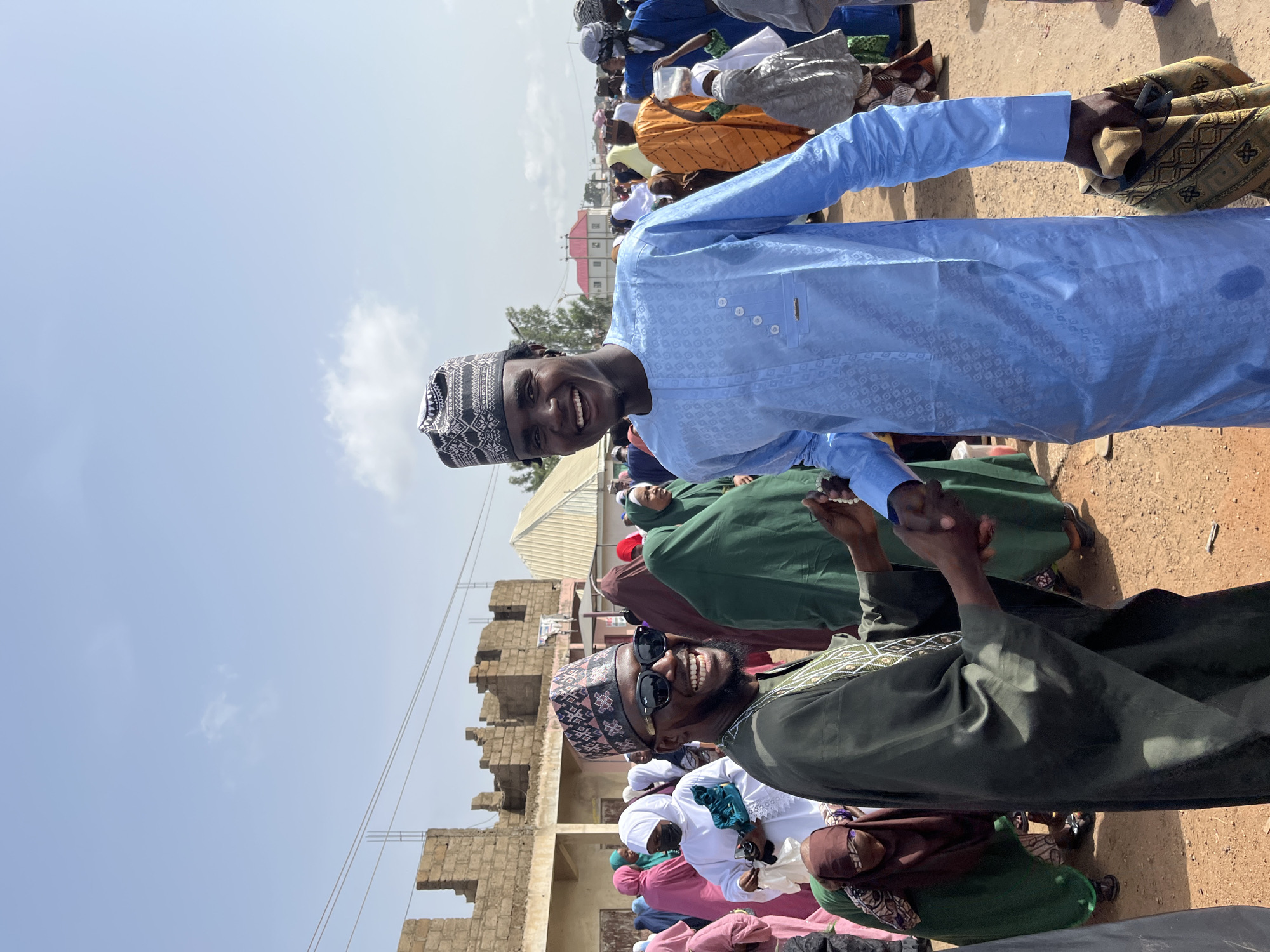 The image shows two men shaking hands in Jos, Nigeria. The man on the left wears a green robe and sunglasses, while the man on the right wears a blue robe. Both men have patterned caps on their heads. They are in a crowded area with other people around them, some wearing traditional clothing and hijabs. A building under construction is visible in the background. The ground appears to be dirt or sand. The sky is light blue with some clouds.