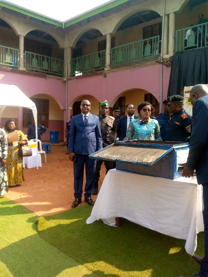 The photo shows a group of people standing in a courtyard. The people are standing in front of a table draped in a white tablecloth. On the table is a blue metal box with writing on it. The people are all wearing formal clothing. Some have military attire. The background shows a pink building with arched doorways and green balconies. The ground is dirt, and there is artificial turf at the bottom of the photo. The photo appears to have been taken in Beni, Democratic Republic of Congo. The coordinates provided in the image description are: 0.491, 29.483.