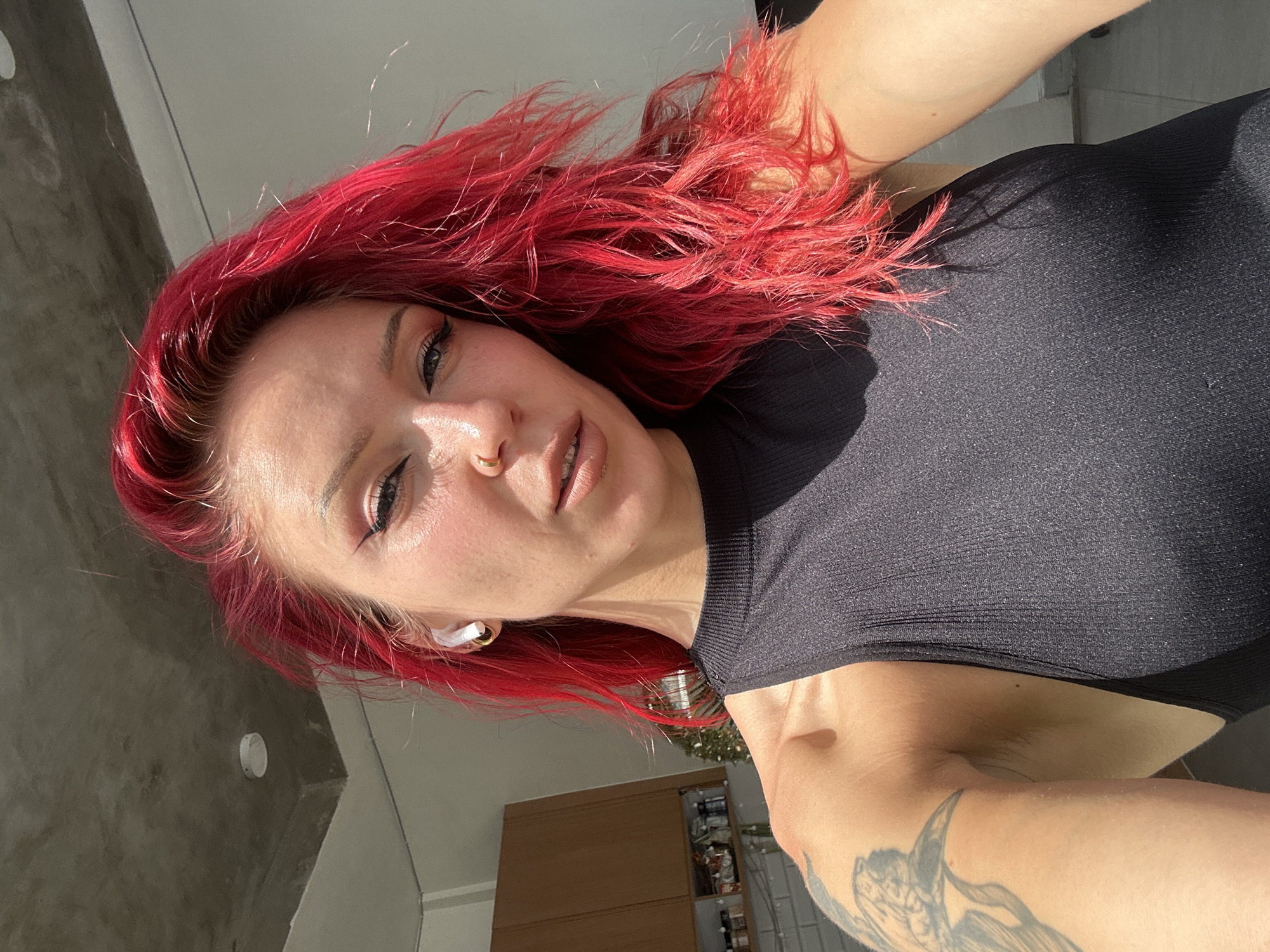 A woman with red hair, a nose ring, and a black top is indoors. She has a tattoo on her arm. There is a cabinet, possibly in a kitchen, and concrete walls. The location is assumed based on inference.