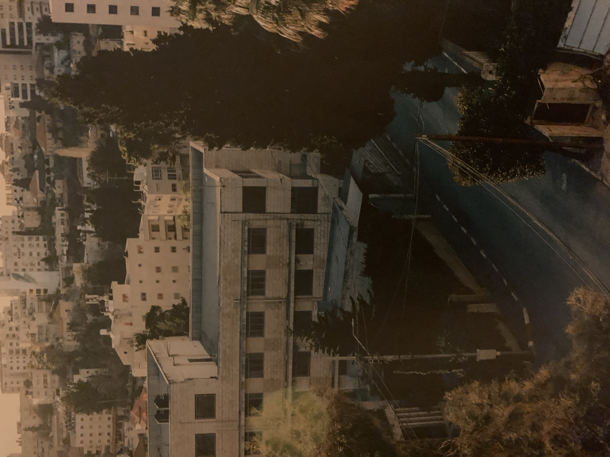 A high-angle, partially obscured view of a dense urban environment likely situated in Barcelona, Spain. Buildings of varying heights and designs constitute the cityscape; primarily constructed in white or light-colored material with rectangular window patterns. Trees and foliage partially obscure the view, suggesting a residential area with some greenery. A paved road runs through the scene, and its surface appears dark, implying either nighttime or shadow cover. A few utility poles with suspended cables suggest the presence of infrastructural elements for power or communication. The photo suggests a moment in time, capturing elements of both urban design and natural scenery.