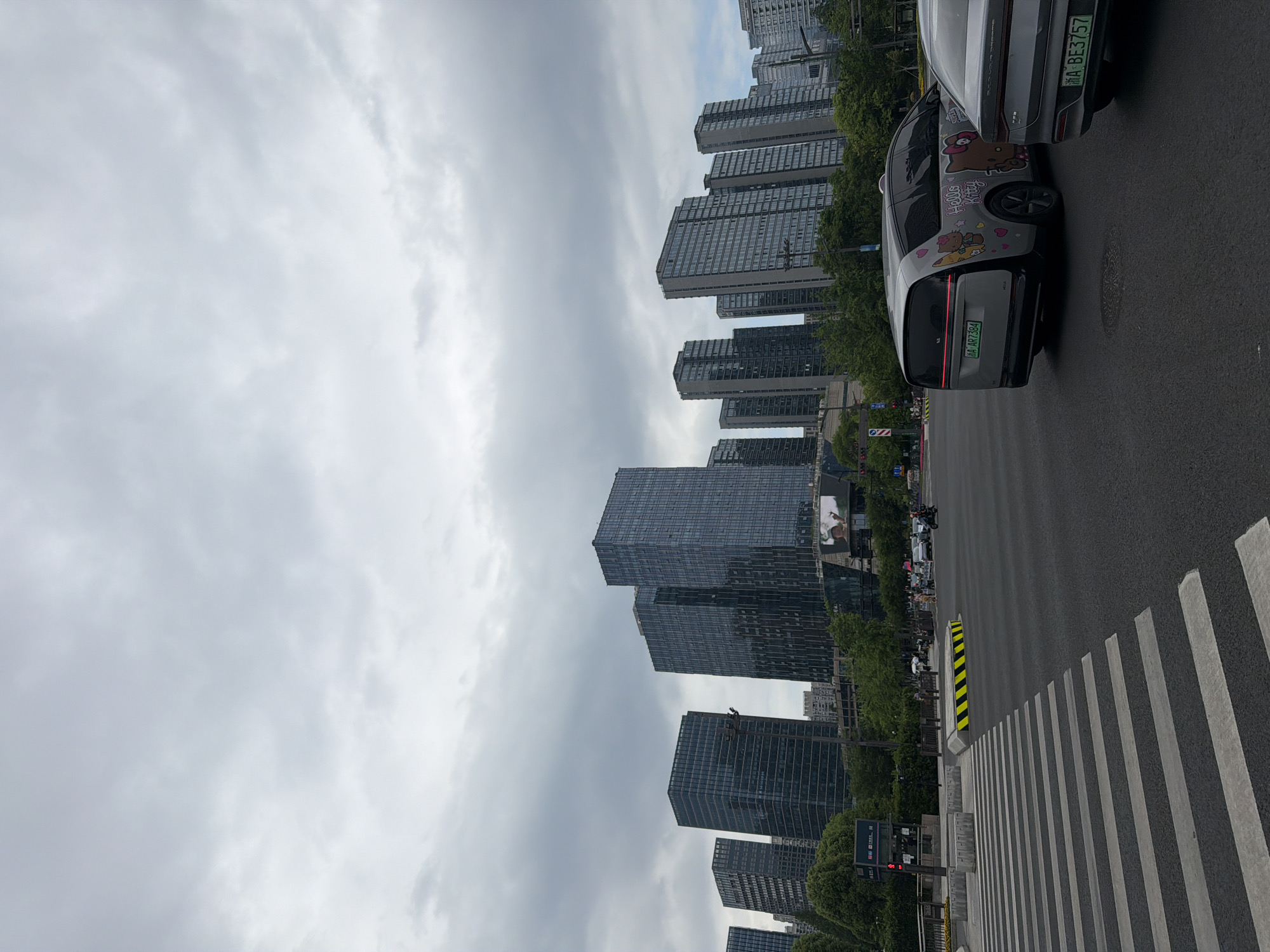 The image shows a street scene in Binjiang District, China. The sky is overcast with heavy clouds. High-rise buildings line the street. Cars are parked on the right side of the road, and a crosswalk is visible. A car decorated with Hello Kitty stickers is among the vehicles. Pedestrians can be seen walking on the sidewalk.