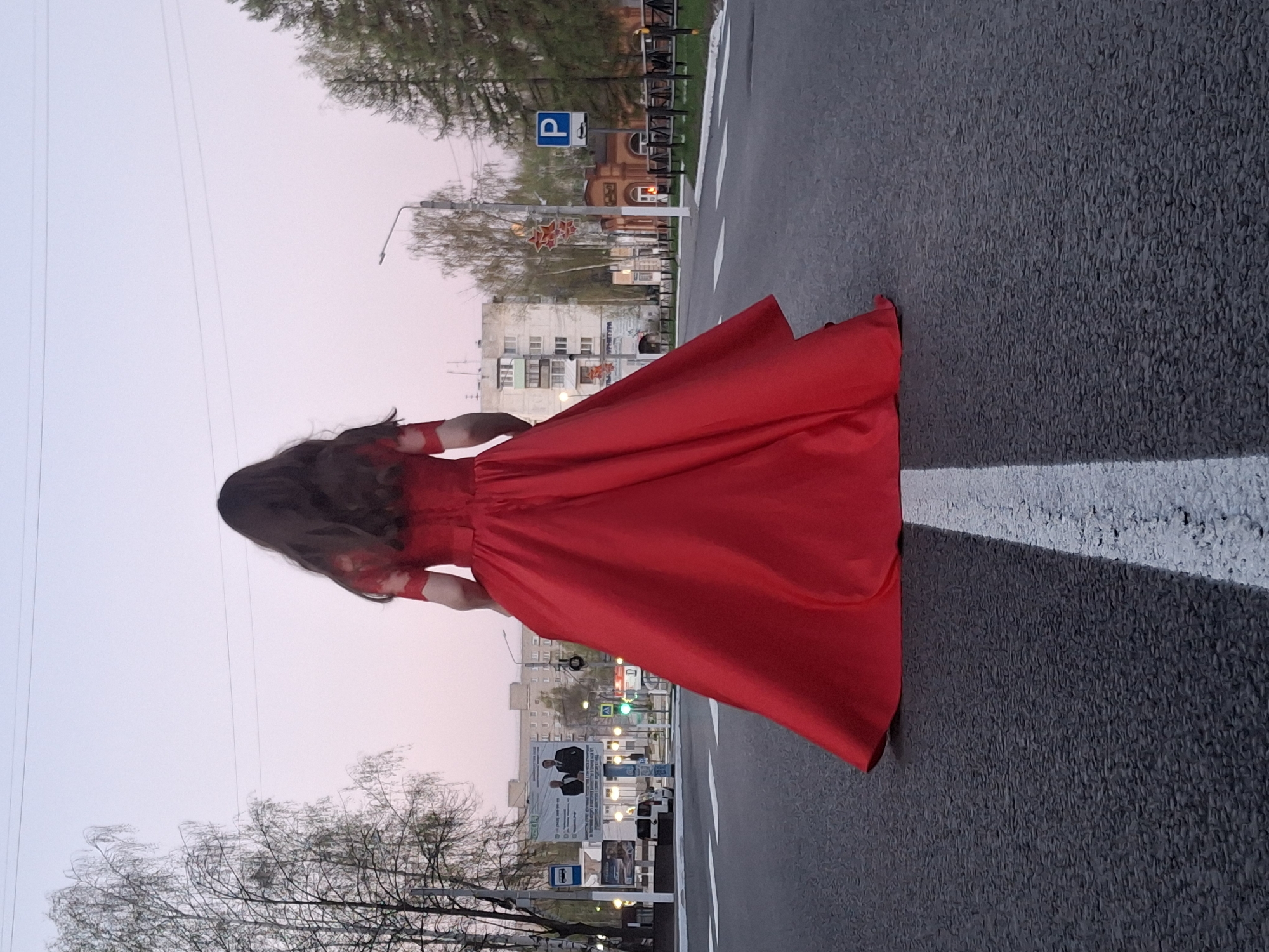 A woman in a flowing red dress stands on a crosswalk in Chaikovsky, Russia. The crosswalk is marked with thick white lines on dark gray asphalt. The woman's back is to the camera, and her long brown hair cascades down her back. A building with a parking sign is visible in the background. Trees line the street, and the sky is a pale pink color, suggesting either sunrise or sunset. A billboard and traffic lights are also visible in the background.