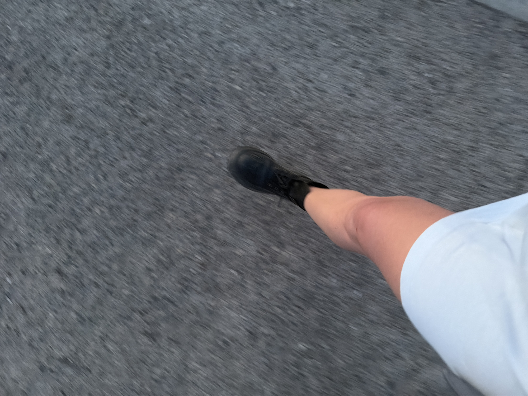The image shows a person walking on a gray asphalt surface. The person is wearing a black boot on their left foot and a white garment is visible around their right leg. The scene is out of focus, suggesting movement. The location, Chaikovsky, Russia, cannot be confirmed from the image.
