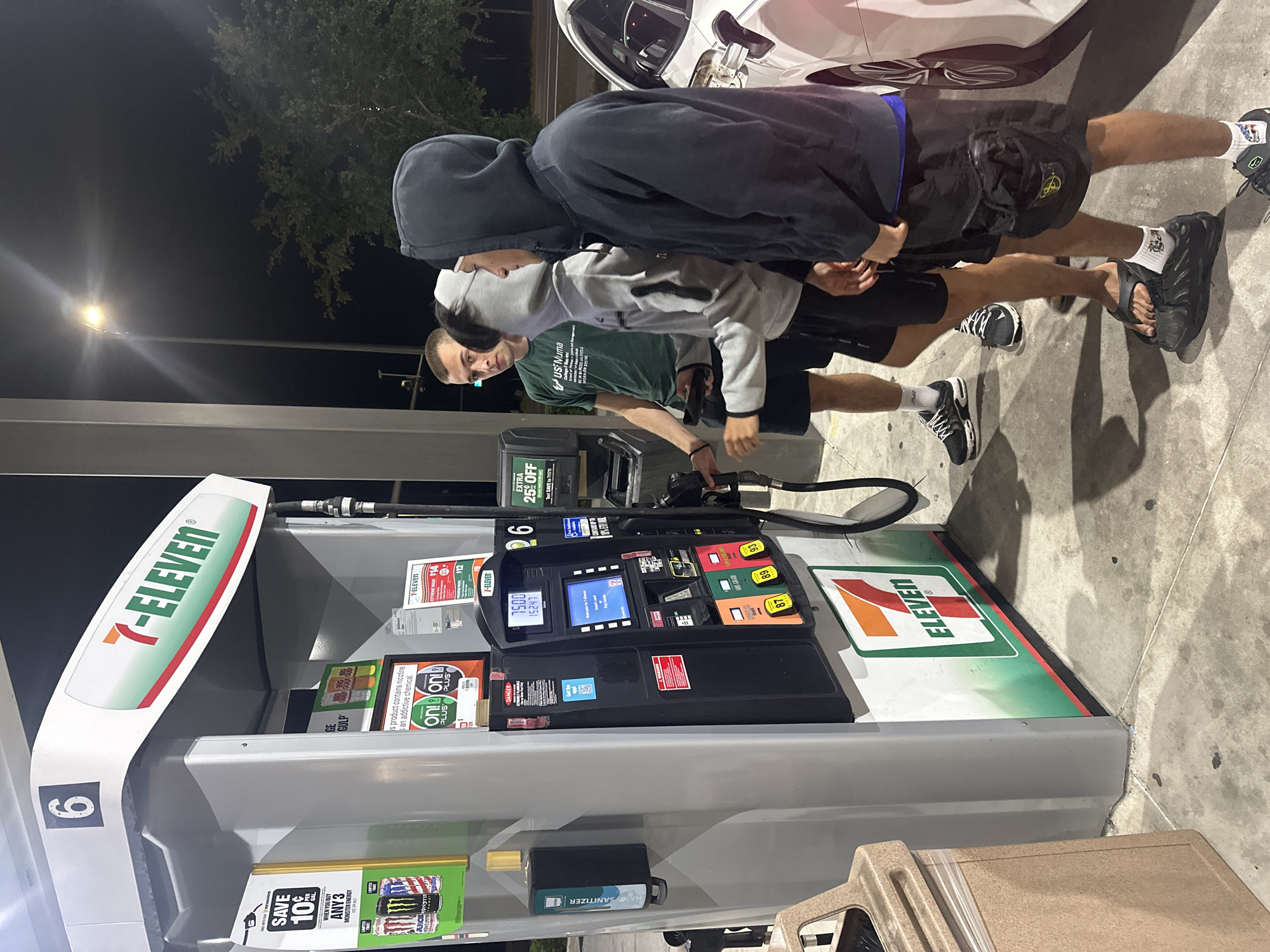 The image shows a scene at a 7-Eleven gas station, with a gas pump displaying $15.00 and 15.247 gallons pumped. Two young males stand near the pump, while another person, possibly filling a white car, is seen. The gas station offers various promotions, including savings on Monster Energy drinks and nicotine products, as well as extra discounts for using the 7-Eleven app. Hand sanitizer is available at the pump. The pump is labeled with the number "6." Based on the "USF Numa" text on a shirt, it is unlikely that the image is in the city of Homosassa Springs, United States of America.