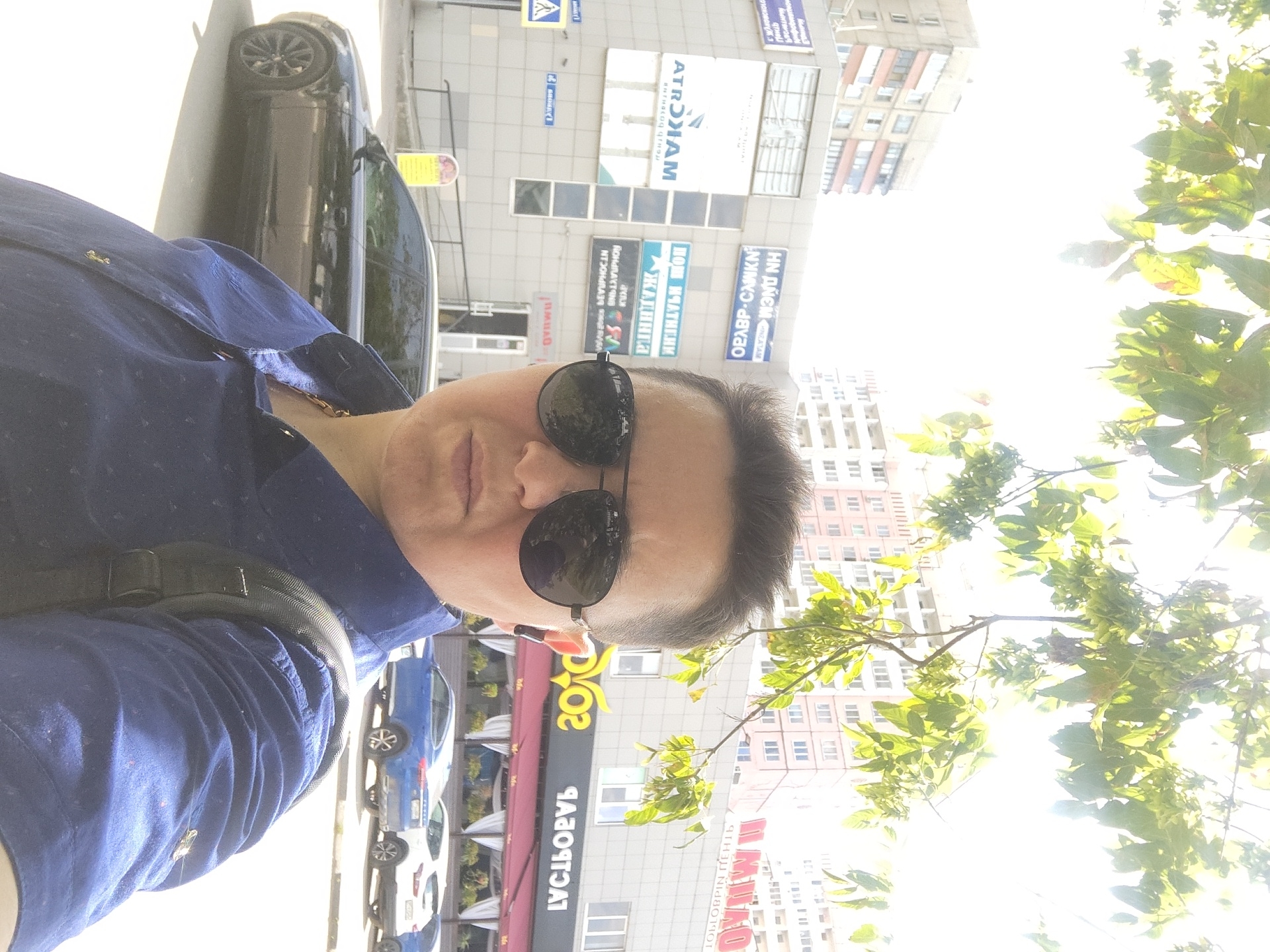 A medium shot captures a person wearing a dark blue collared shirt, a gold chain, and black aviator sunglasses, taking a selfie outdoors. The person has short hair and is positioned slightly off-center to the right. Behind them, a building with several windows and signs is visible. One sign, in blue and white letters, reads "HN ДНЁМ" and another, also in blue and white, displays "АТРОХАМ". Further down the street, a row of parked cars, including a black Audi, a blue crossover SUV, and a white SUV, are present. The background is dominated by apartment buildings and foliage from trees. The sky is bright, indicating a sunny day. The overall scene appears to be a street in the city of Zhukovsky, Russia, likely in front of a commercial building.