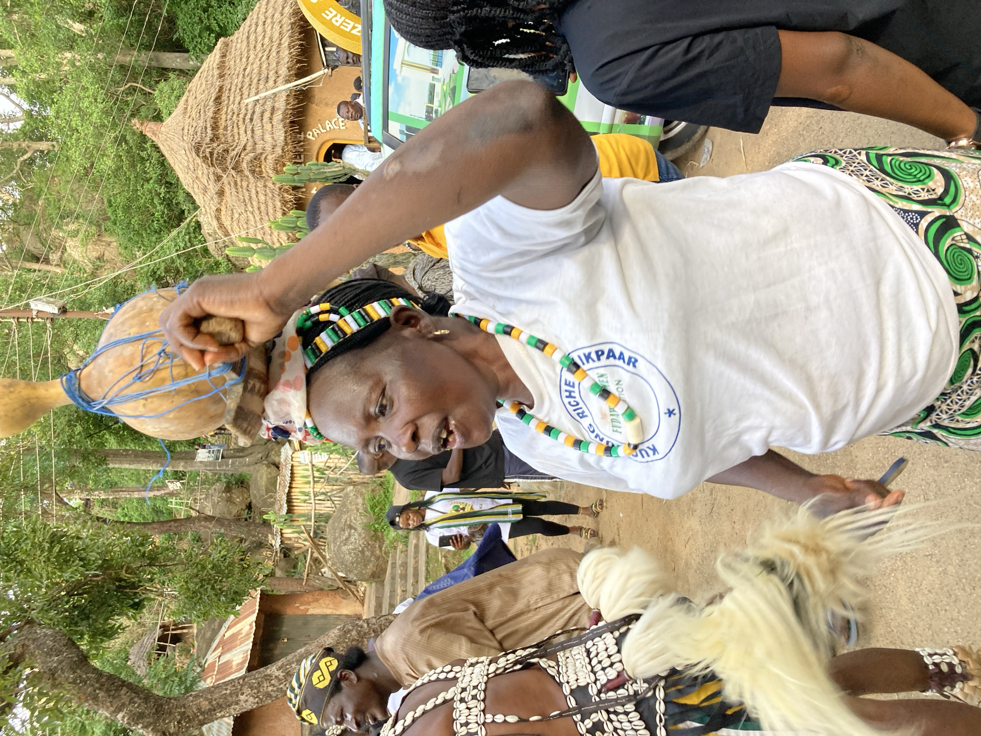 In Jos, Nigeria, a woman is carrying a calabash on her head, secured with blue rope. She wears a white t-shirt with a circular blue logo and a colorful beaded necklace. Her hair is braided and adorned with green, black, and white beads. She is participating in a cultural event, possibly a procession or festival, as indicated by the traditional attire and festive atmosphere. Other individuals in the background are also dressed in traditional clothing. The environment suggests an outdoor setting, possibly a village or a community gathering place, with thatched-roof structures and lush vegetation. The overall scene captures a moment of cultural celebration and community engagement in Jos.