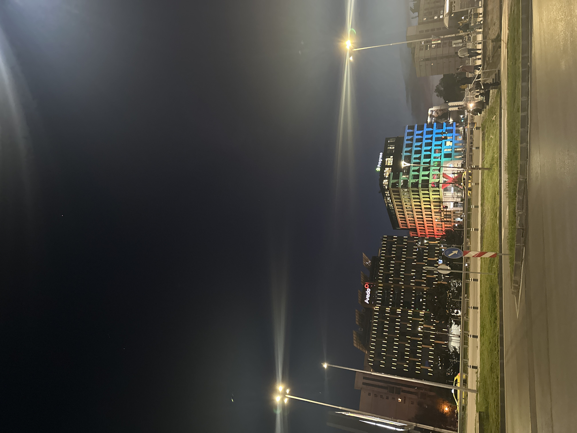 This nighttime photograph captures a modern urban street scene in Sofia, Bulgaria. The dark sky is illuminated by artificial lights, creating a vibrant city atmosphere.
On the left side of the frame, two multi-story buildings are prominently featured. The upper building displays a striking, colorful facade, illuminated with a gradient of rainbow hues, transitioning from blues and greens at its top to yellows, oranges, and reds at its lower sections. The word "Progress" is clearly visible on this building. The lower building is adorned with numerous warm-toned, rectangular lights arranged in a grid pattern across its exterior. The text "Avedo O" (likely a logo) is discernible on its facade.
The right side of the image depicts a multi-lane road, which appears reflective under the artificial light. A narrow grassy median separates the road from the buildings. Several streetlights emit bright white light, casting noticeable lens flares across the dark background.
A few blurred figures, possibly pedestrians, are visible near the colorful building, indicating some human activity. One individual appears to be on a scooter or motorcycle near the median strip. A blue circular road sign with a white right-pointing arrow and a red and white striped barrier are also present near the lower building. The scene conveys a sense of a contemporary city environment after dark, characterized by dynamic architectural lighting.