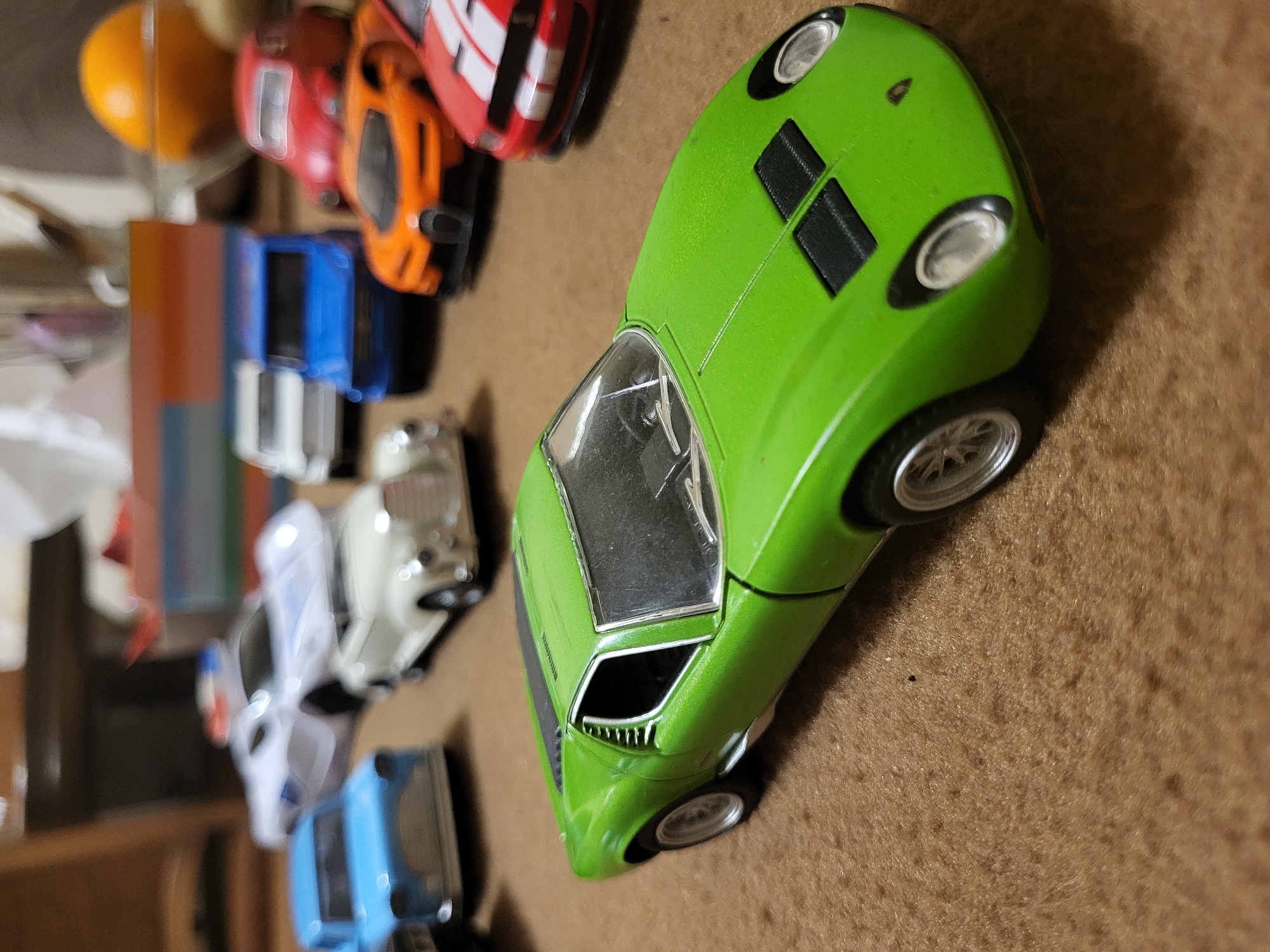The image shows a collection of toy cars arranged on a brown surface. The cars appear to be die-cast models of various makes and colors. Prominent among them is a green car, possibly a Lamborghini Miura, positioned towards the right side of the frame. Several other toy cars, including red, orange, blue, and white models, are lined up behind it, leading towards the left edge of the image. An orange ball is located at the top left of the image. There is no information about the city of Yerevan, Armenia, in the picture.