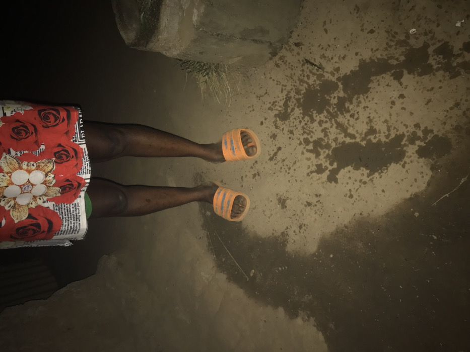 A person in orange sandals stands on a wet, muddy surface. A concrete or stone structure is in the upper left corner. The person wears a skirt or wrap with a red floral pattern and newspaper print. The surrounding area is dark, with the ground appearing muddy and uneven. The picture is taken from an overhead perspective.