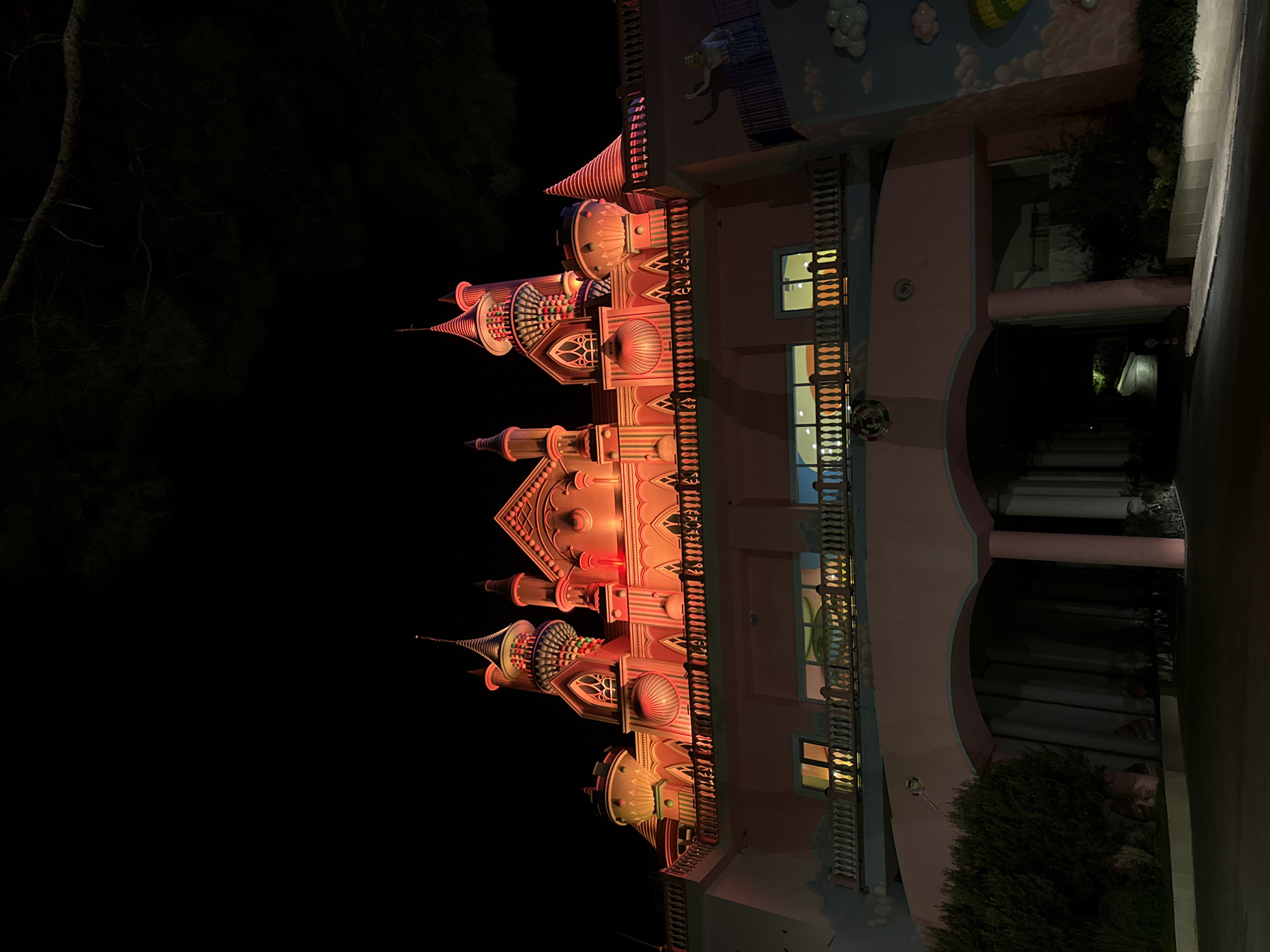 The image shows a brightly lit, pink castle-like building at night in Bodrum, Turkey. The building has multiple towers and ornate architectural details. Red lights illuminate the upper sections of the castle, creating a dramatic effect. A tree branch extends into the frame from the top left, silhouetted against the dark night sky. In the foreground, there is a dark paved area, likely a road or driveway, leading to arched entrances at the base of the building. There are some bushes and greenery visible on the left side of the image. The overall impression is of a whimsical or entertainment-oriented structure, possibly a theme park or a hotel.