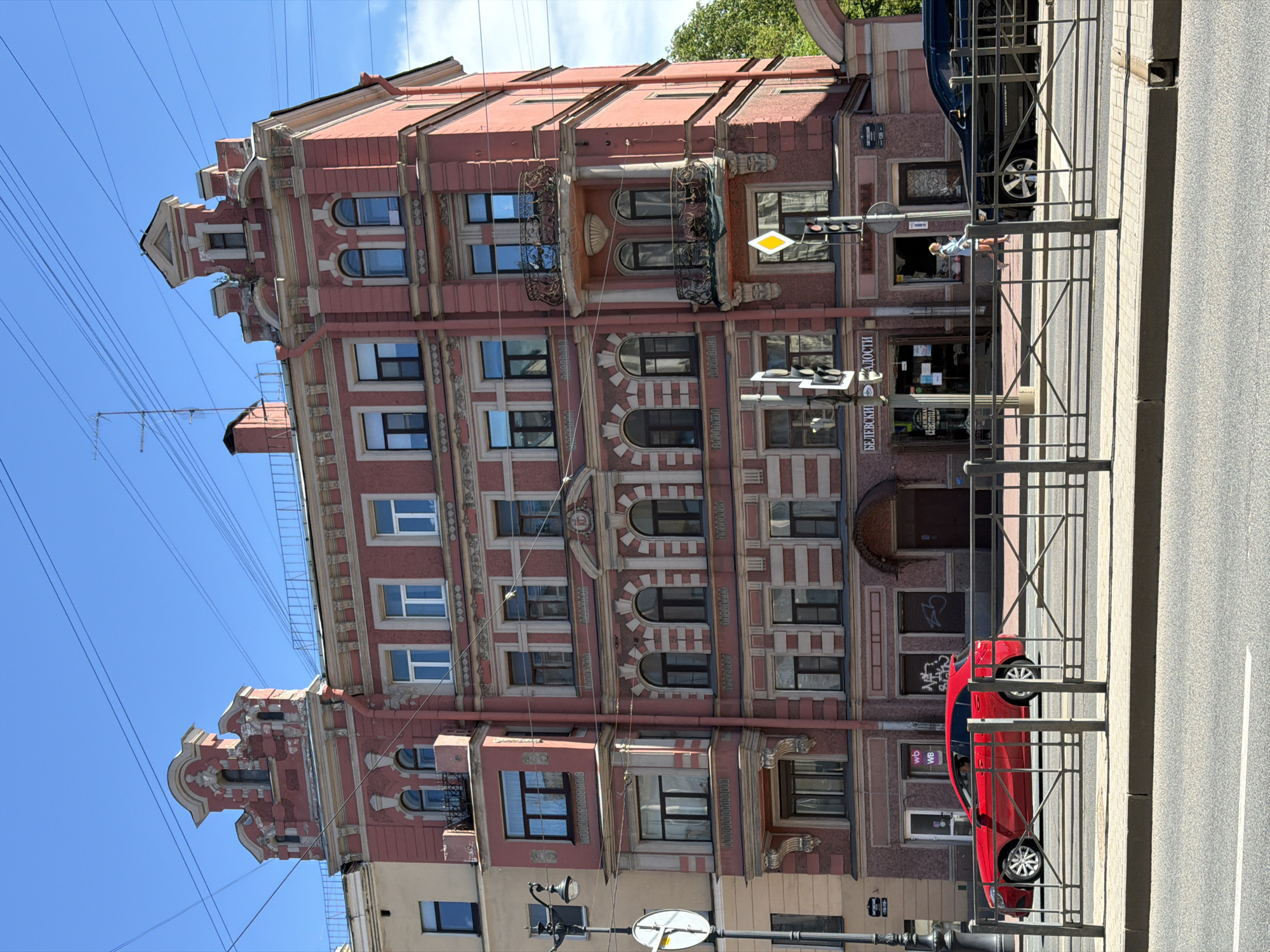 This image captures a daytime street scene on Ligovsky Prospekt, Saint Petersburg, Russia. The central focus is a six-story (including decorative roof features) building painted in a salmon-pink hue, exhibiting an eclectic architectural style with ornate detailing.
The building's facade is adorned with white pilasters, cornices, and decorative bands. It features numerous windows, some rectangular and some arched, predominantly with white frames, though some have darker frames. Multiple balconies with intricate black wrought-iron railings are visible on various floors. The roofline is punctuated by two prominent, ornate pediments at the corners; the left one shows visible paint wear, while the right one hosts a small rooftop antenna. The ground floor houses commercial establishments, including a storefront on the left displaying a "wb" logo and a central shop with Cyrillic signage that reads "БЕЛЕВСКИЕ СЛАДОСТИ" (Belevskie Sweets) and "ПОЧТА РОССИИ" (Russian Post). A lighter, yellowish building is partially visible adjacent to the main structure on its left.
In the foreground, an asphalt road with white lane markings runs parallel to a concrete sidewalk, separated by a dark metal barrier fence with an 'X' pattern. Overhead utility lines are prominent, crisscrossing the clear blue sky. Two black lampposts are present, one on the left and one on the right, both supporting traffic signals that include yellow diamond-shaped 'priority road' signs.
A single pedestrian, a woman dressed in a light blue top and a white skirt, is walking on the right side of the sidewalk. Two vehicles are on the road, partially obscured by the foreground fence: a red hatchback car on the left and a black SUV on the right. Green foliage from trees is visible in the upper right background.