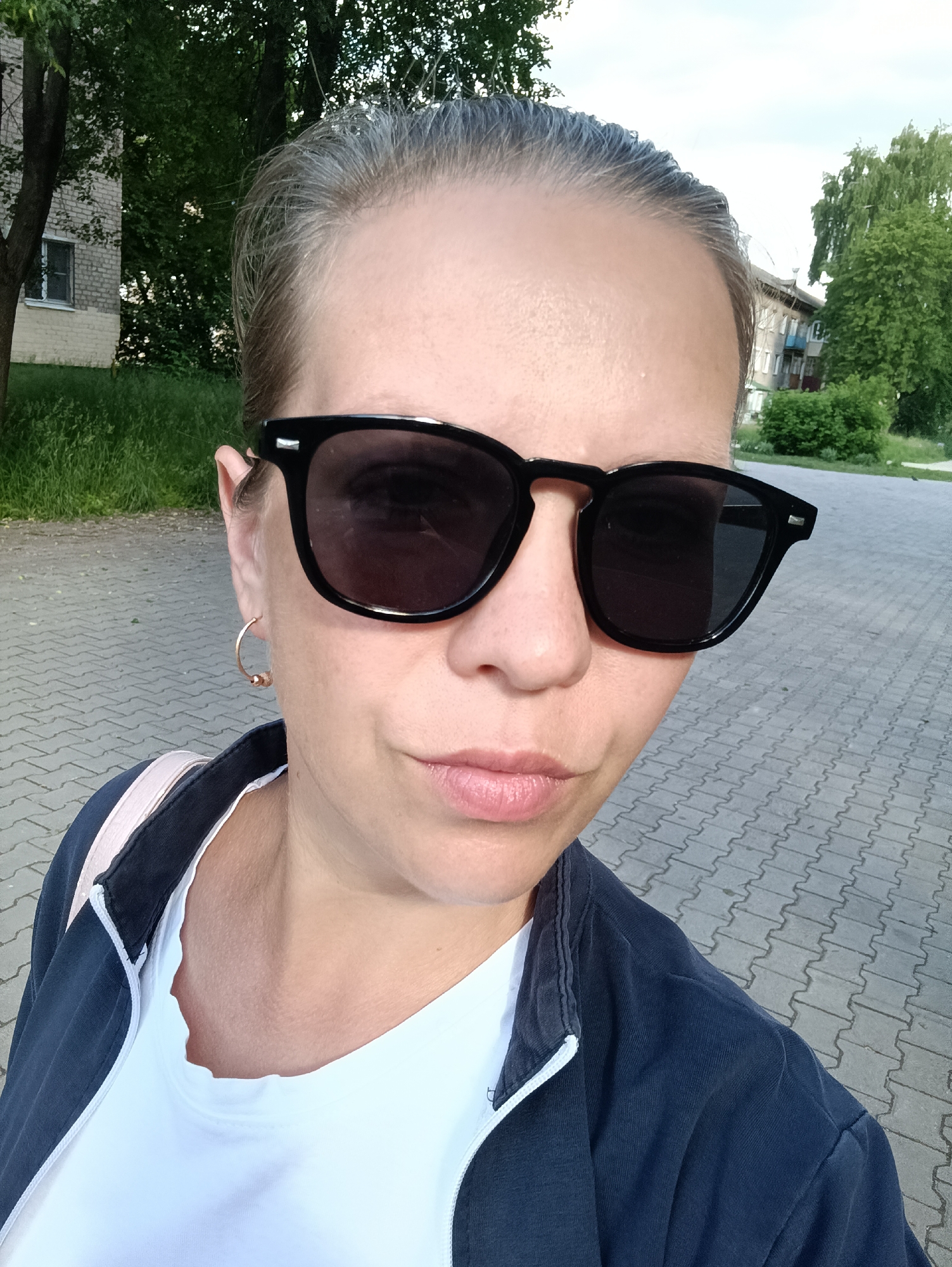 The image depicts an adult female engaged in a self-portrait, or being photographed, outdoors in what appears to be a residential area of the city of Chaikovsky, Russia. The woman, positioned centrally and looking towards the camera, has dark blonde or light brown hair pulled back, with visible graying at the hairline. She wears dark-lensed, black-framed sunglasses that obscure her eyes. Her skin appears light-toned, and her lips are closed with a subtle pink hue. A small, gold-colored hoop earring with a bead is visible on her left ear. She is dressed in a white t-shirt underneath a dark blue or navy zippered jacket or sweatshirt. A light pink strap, possibly from a bag, is visible over her left shoulder.
The environment consists of a paved ground surface made of interlocking light gray concrete pavers, forming a patterned walkway or courtyard. The background includes green foliage and trees, characteristic of a leafy urban or suburban setting. To the left, a light-colored brick building with a visible window is partially obscured by trees. To the right, further in the distance, another multi-story residential building with balconies is visible. The sky above is overcast, indicating daylight conditions with diffused light.