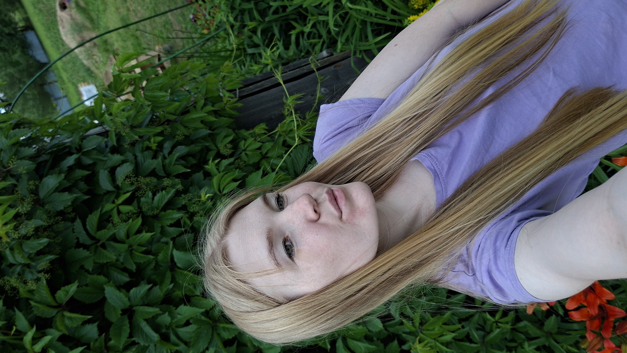 A fair-skinned young woman with long blonde hair is taking a selfie outdoors in the daytime. She is wearing a light purple t-shirt and is positioned in the foreground. Behind her, a dense wall of green foliage, likely ivy, covers a wooden structure. To the right of the foliage, there is a glimpse of a grassy area with some scattered objects, including what appears to be a garden bed and a mound of earth. The overall setting suggests a garden or yard in a rural or suburban environment.
