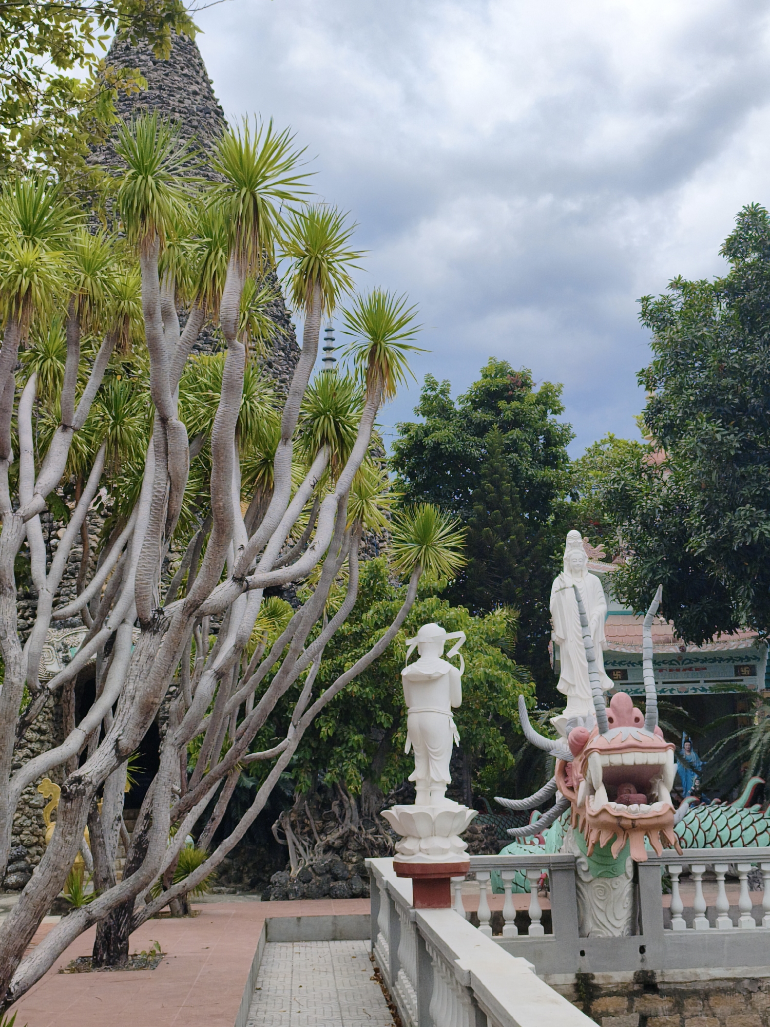 The image captures an outdoor scene at a Buddhist temple in Cam Ranh, Vietnam. In the foreground, a white statue of a standing figure, possibly a deity, is placed on a lotus pedestal. Adjacent to it, a vibrant pink and green dragon sculpture with its mouth open and teeth bared is a prominent feature, appearing to emerge from a decorative railing. Further back, another taller, serene white statue of a standing figure is visible. The surrounding environment is lush with vegetation, featuring a cluster of unique trees with thick, grey, branching trunks and spiky green fronds. Behind the trees, a tall, conical stone structure, possibly a stupa, can be seen, with a distant pagoda-like tower peeking through the foliage. The sky above is cloudy and overcast. The scene is a blend of religious iconography, natural elements, and architectural structures typical of a Buddhist temple complex.