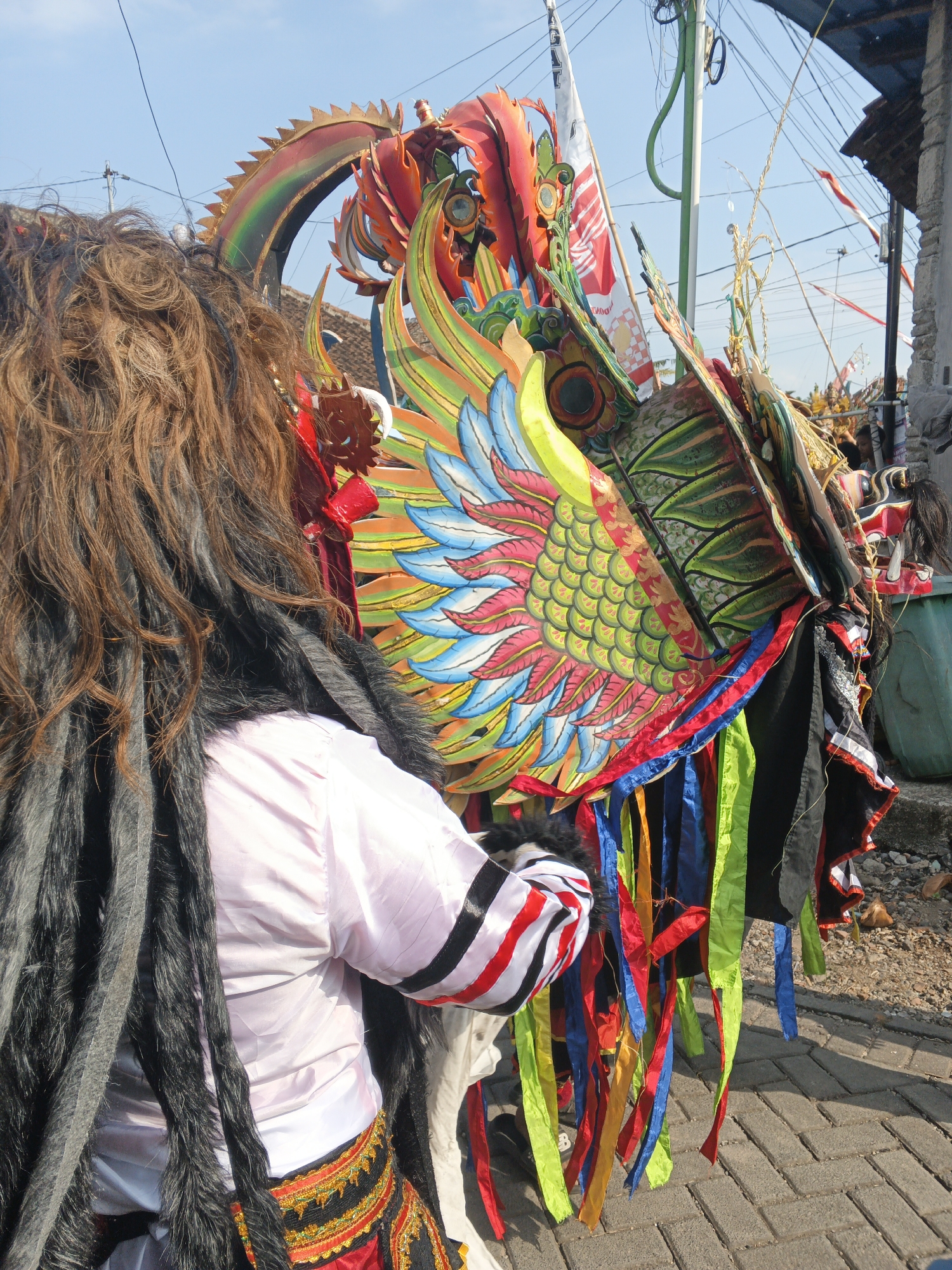 In Singolatren, Indonesia, a person dressed in a costume, possibly for a parade or festival, is seen from the back. The costume features a vibrant, intricately designed dragon-like headdress adorned with bright colors such as red, orange, yellow, green, and blue, with feather-like elements and scale patterns. The wearer has long, brown, curly hair and is wearing a pink shirt with black and red striped cuffs. A black, furry trim is visible around the neck and shoulders of the costume. The lower part of the costume includes a red skirt or fabric with an ornate, embroidered waistband in yellow, black, and red. Colorful streamers in red, blue, green, and yellow hang from the costume, adding to its festive appearance. The scene takes place outdoors on a paved area, possibly a street, with a clear blue sky overhead. In the background, there are hints of buildings and utility poles with wires, suggesting an urban or semi-urban setting within the city. Other decorative elements, like flags and more costume pieces, are visible in the distance, indicating a communal event is underway.