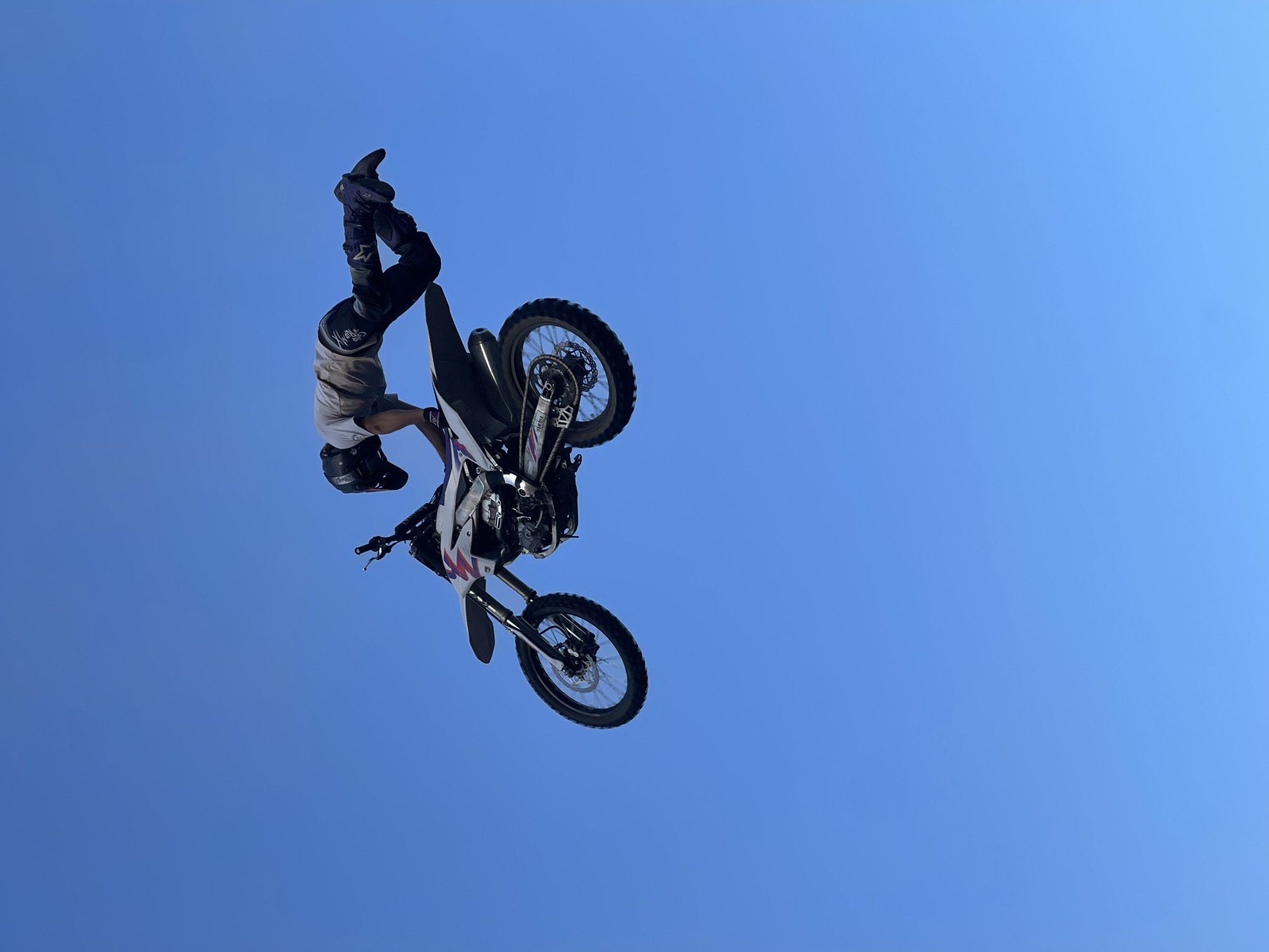 In the city of Strathroy-Caradoc, Canada, a motocross rider performs a stunt against a clear blue sky. The rider, wearing a helmet, light-colored t-shirt, and dark pants with knee pads, is suspended mid-air, with their body arched backward over a dirt bike. The motorcycle, predominantly white with red and blue accents, is also airborne, positioned beneath the rider. The front wheel of the bike is tilted upwards, while the rear wheel is angled downwards, suggesting a dynamic maneuver. The scene is devoid of any visible background elements or structures, focusing entirely on the aerial display of the rider and their motorcycle against the vast expanse of the sky.