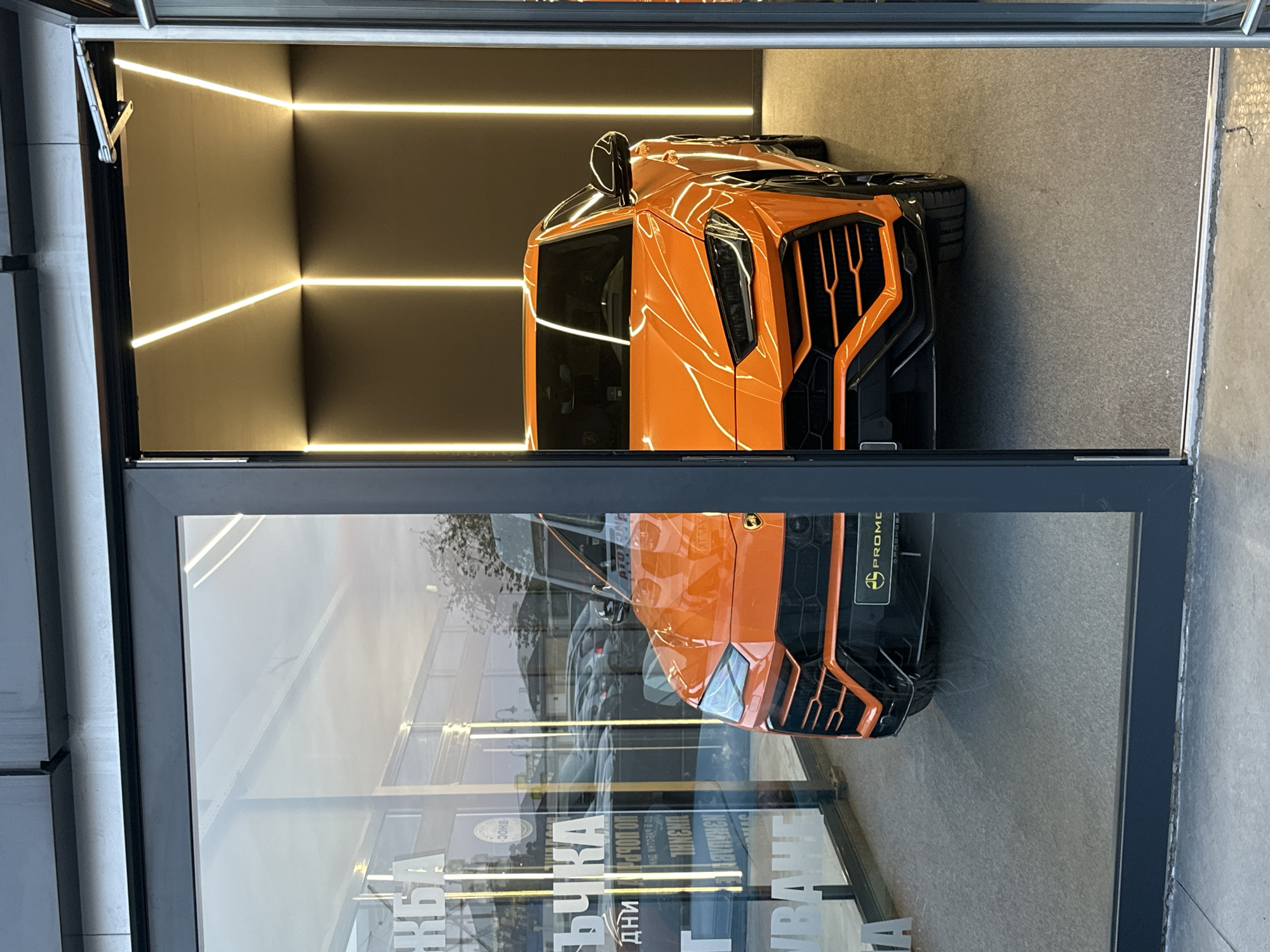 The image shows an orange Lamborghini Urus Novitec Esteso SUV on display in what appears to be a storefront. The car is viewed through the shop's window, and a reflection of the surrounding environment, including street signs and possibly other buildings, is visible on the glass. The shop's interior has a modern design, with dark gray walls and horizontal LED strip lighting. The front of the car is prominently featured, including its distinctive grille and headlights. The location is likely in Varna, Bulgaria, as the window display appears to have a text that includes Cyrillic letters, and the car is on display in a storefront.