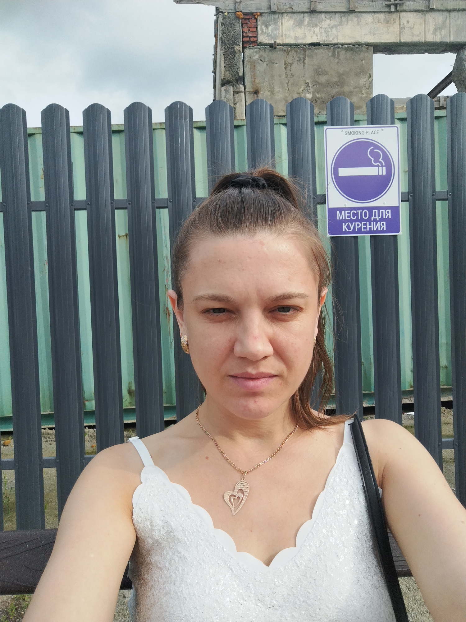 In an outdoor, urban setting, a woman with her hair pulled into a ponytail takes a selfie. She is wearing a white, sequined top and a gold necklace with a heart-shaped pendant. She is positioned in front of a dark gray metal fence, which runs horizontally across the frame. Behind the fence, a light green structure is visible, and further in the background, the remains of a building with visible brickwork and concrete structures suggest an industrial or derelict area. To the right of the woman, a white sign with a purple border is attached to the fence, depicting a cigarette with smoke and the text "SMOKING PLACE" and "МЕСТО ДЛЯ КУРЕНИЯ" in English and Russian, respectively, indicating a designated smoking area. The sky above is overcast.