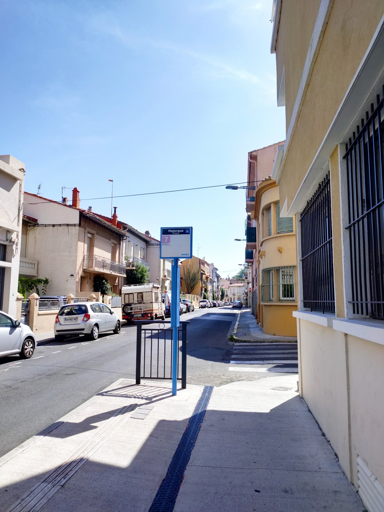 The image depicts a street scene in Perpignan, France, identified by a bus stop sign displaying "Majorque" and "Sanéo," which are local transportation terms in Perpignan. The scene is captured on a clear, sunny day, with a bright blue sky overhead and distinct shadows cast by buildings and objects.
On the left side of the street, a row of residential buildings with beige and light pink facades lines the road. A white car is parked parallel to the curb, and another silver car is visible further down the street. The sidewalk is paved with concrete and features tactile paving for the visually impaired. A black metal gate stands near the sidewalk.
The street itself is paved with asphalt and has white lane markings. Several cars are parked along the right side of the street. A white recreational vehicle or camper is parked on the left side, in front of a building. Further down the street, more buildings are visible, creating a sense of an urban or suburban environment.
A prominent blue bus stop sign stands on the right side of the street. It displays the number "7" in a red circle, indicating a bus route, and the text "Majorque" and "Paris des Sports." The sign also features a blue accessibility icon. Across the street from the bus stop, a building with a yellow facade and dark metal bars on its windows is prominent. A pedestrian crossing is marked with white stripes on the asphalt.
The overall impression is of a quiet, residential street on a bright day.