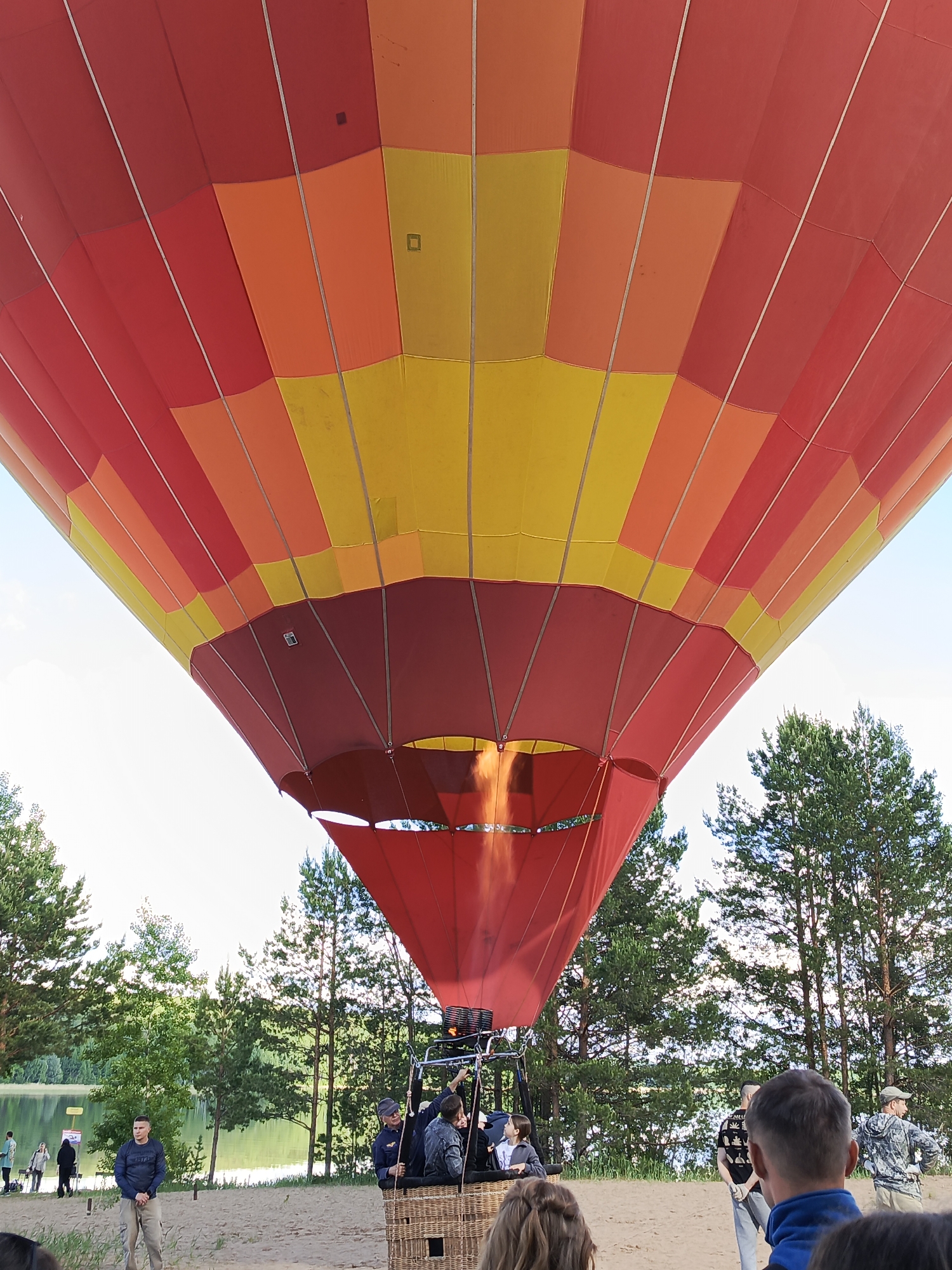 A colorful hot air balloon, primarily red, orange, and yellow, is being inflated or prepped for flight. The balloon's fabric is panelled, with visible flame emanating from the burner area beneath. Below the balloon, a wicker basket contains several people, possibly pilots or passengers. A group of onlookers surrounds the balloon, some of whom are visible in the foreground and background. In the background, there is a lake and a line of trees. The scene is set on a sandy ground, potentially a beach or clearing near the water. The image was taken in Chaykovsky, Russia.