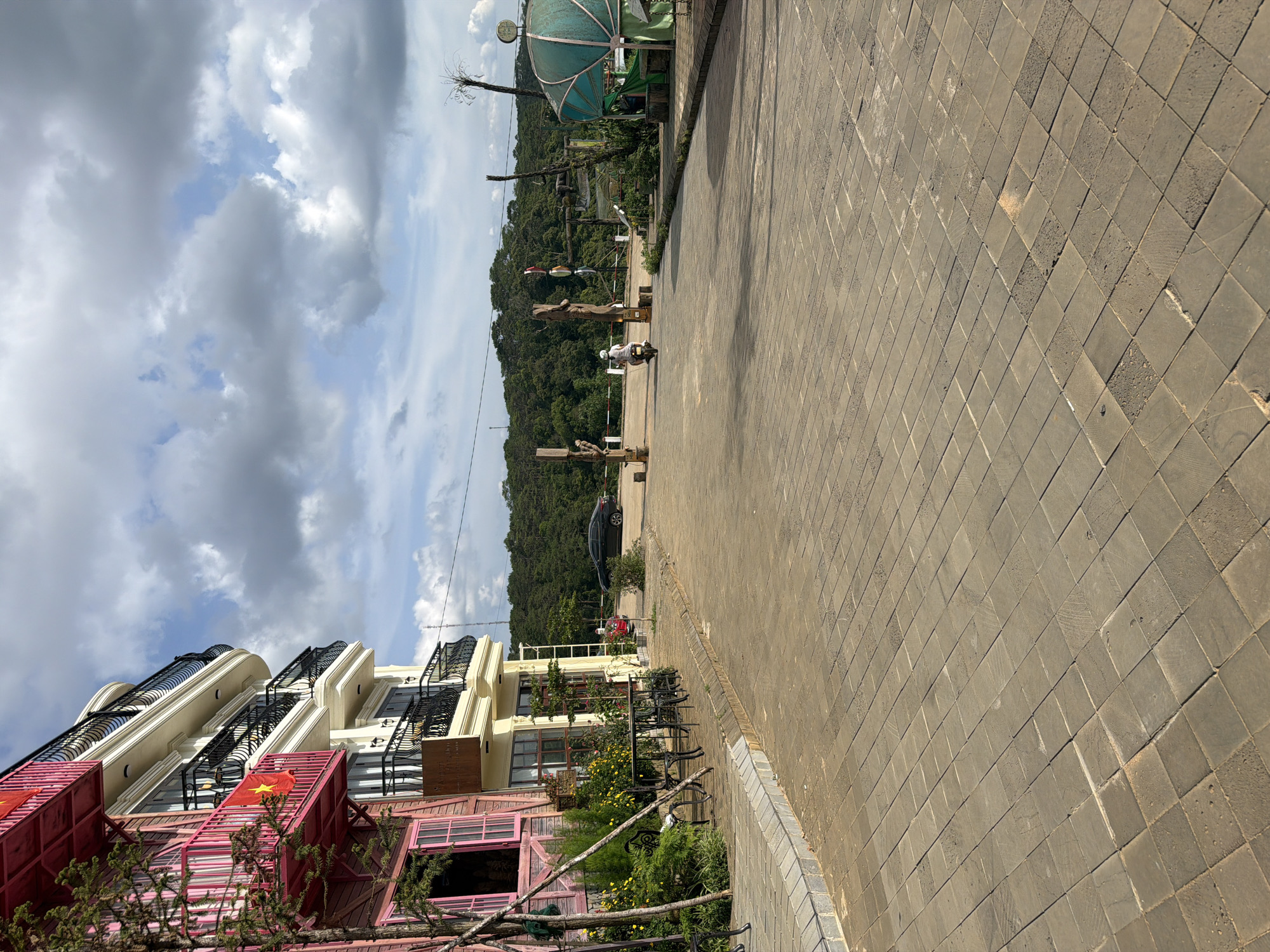 The image displays an outdoor street scene in Măng Đen, Vietnam, under a bright, partly cloudy sky. The perspective is taken from a wide, tiled pavement on the right, looking down a paved road that extends into the distance.
On the left side of the road, a row of multi-story buildings is visible. The lower sections of these buildings feature striking pink and red corrugated metal facades, adorned with several Vietnamese flags (red with a yellow star). Above, the buildings transition to cream-colored exteriors with elegant black, ornate balcony railings. In front of these structures, a small garden area bursts with green plants and bright yellow flowers, alongside outdoor tables and chairs, suggesting a cafe or restaurant.
Along the paved road, a person is seen riding a white scooter, heading away from the viewer. Further down, a black car and a red car are parked. The roadside is decorated with large, rustic wooden posts, some of which appear to have lanterns or other embellishments. In the mid-ground, a distinctive light green or teal-colored gazebo-like structure stands out.
The background is dominated by a vast expanse of densely forested hills, indicating a mountainous or hilly setting. The sky overhead is a mix of light blue patches and large, fluffy grey and white clouds. The lighting suggests it is daytime, casting clear shadows on the pavement. The scene combines urban or commercial development with a rich natural landscape. No readable text is visible other than the emblem on the Vietnamese flags.