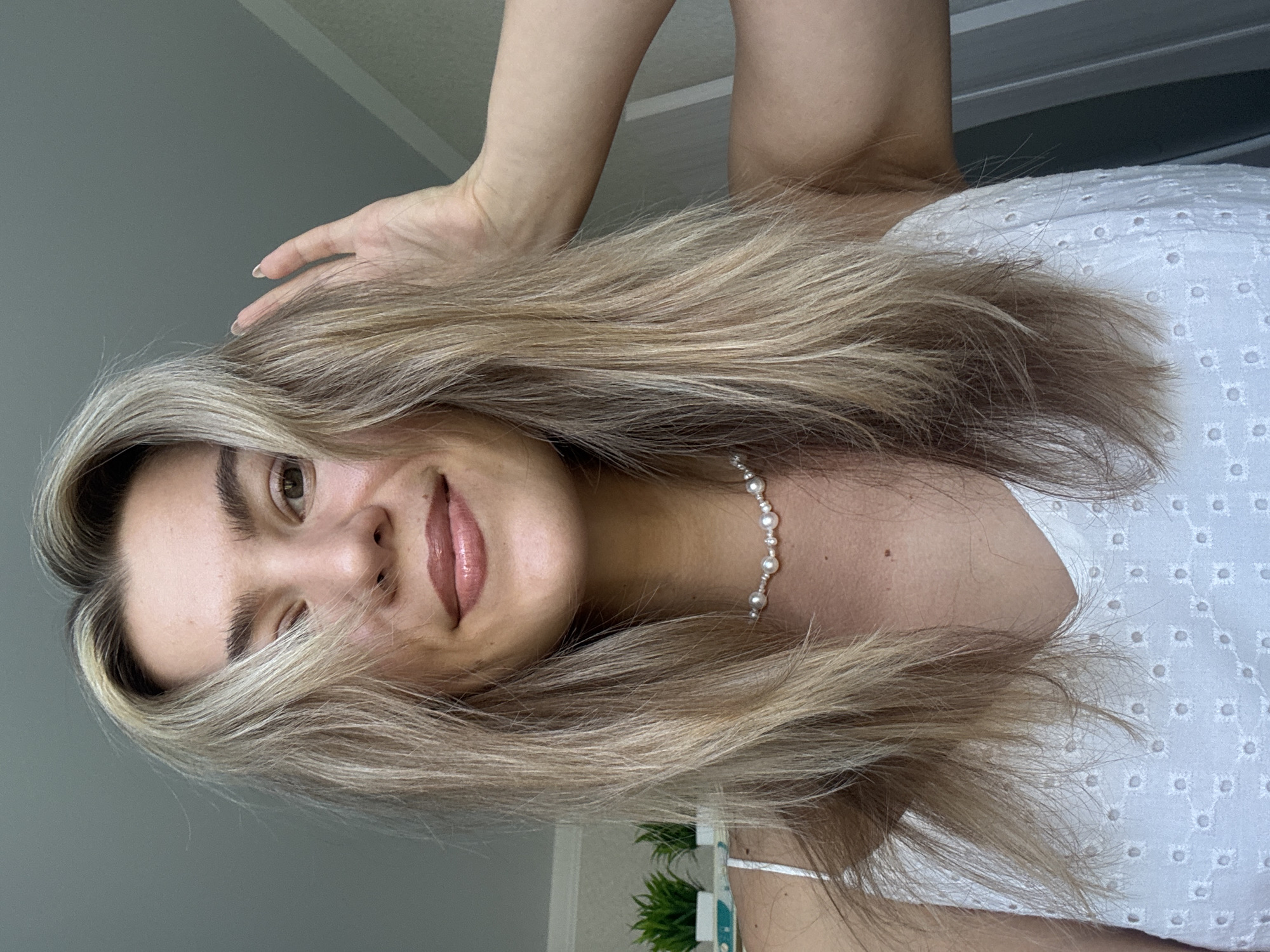 A woman is shown from the shoulders up, with her head tilted. Her hair is long, blond, and styled in waves. She is wearing a white dress with eyelet detailing and a pearl necklace. Her hand is raised towards her hair. The background includes a light gray wall, a white door frame, and a glimpse of a green plant. The location appears to be indoors.